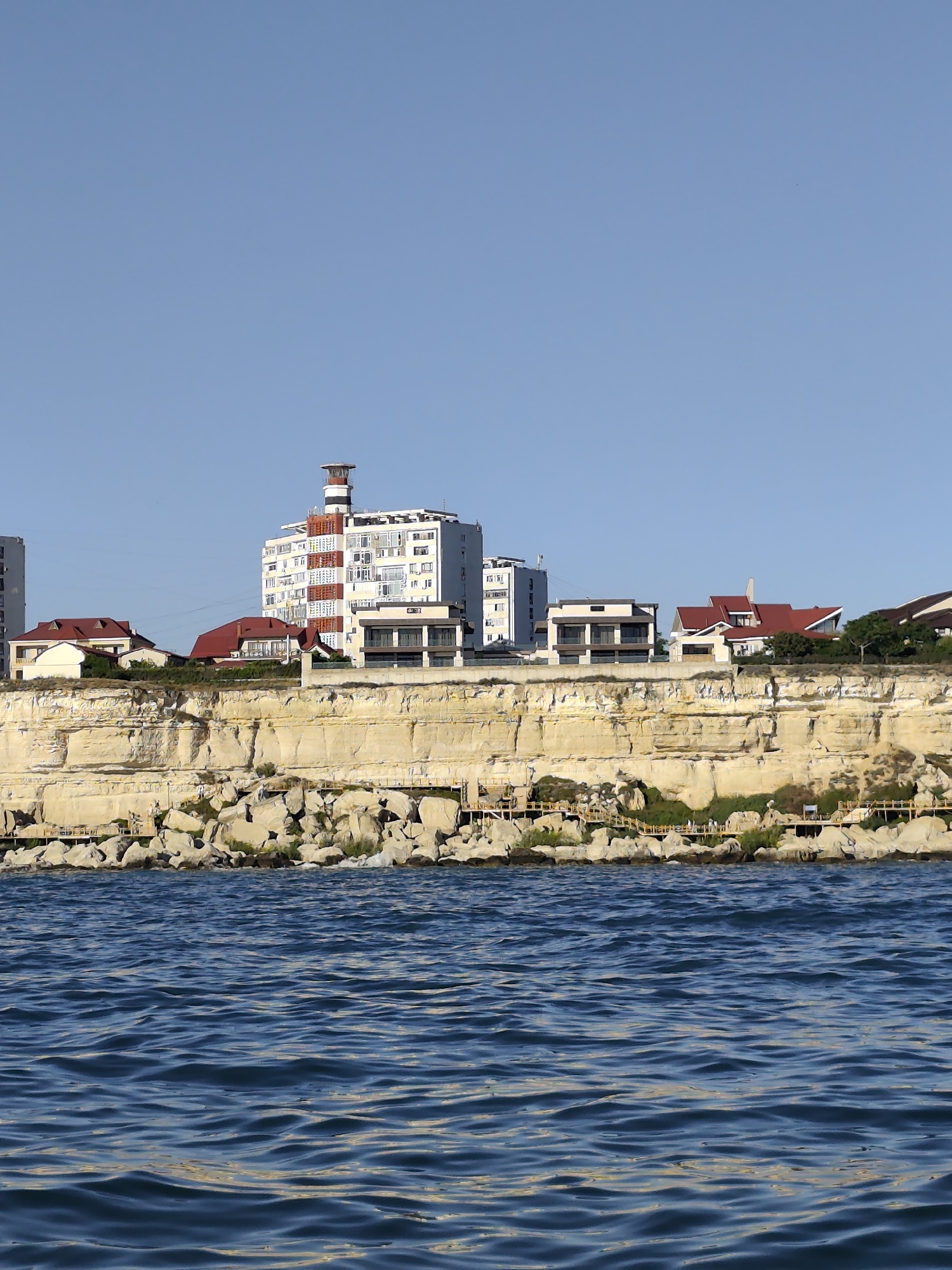 The image shows a body of water in the foreground, with a cliff face and buildings along the top of the cliff in the background. The water is blue and appears to be slightly choppy. The cliff face is a pale yellow color and is covered in rocks. There are several buildings on top of the cliff, including a tall white building with red and white stripes, and several smaller houses with red roofs. There are also some trees and vegetation on the cliff. The sky is clear and blue. The scene appears to be a coastal town.