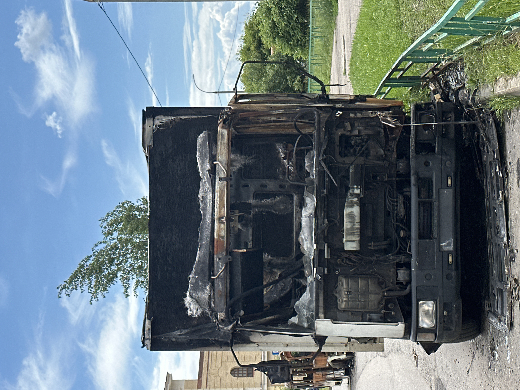 This image shows the front of a severely burned truck, likely a commercial vehicle, in Podolsk, Russia. The truck's cab and engine compartment are heavily damaged, with exposed metal, melted components, and charred debris. The hood is open, revealing the extent of the fire damage to the engine. The headlights and front grille are still somewhat intact, though also scorched.
The truck is parked on an asphalt road next to a grassy area and a sidewalk. A green metal fence runs along the edge of the grassy area. In the background, there are trees and a building, suggesting an urban or suburban setting. The sky is blue with scattered white clouds, indicating it is daytime. The overall impression is one of destruction and the aftermath of a significant fire. There is no visible text in the image. The scene appears to be set on a clear, sunny day.