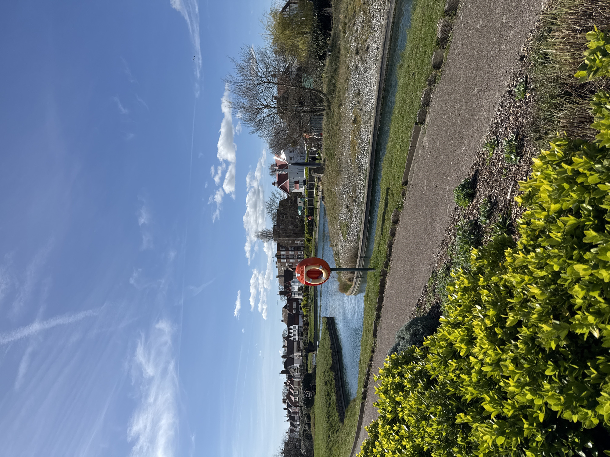 The image shows a canal in Great Yarmouth, United Kingdom. The canal is lined with grassy banks and stone walls. A narrow paved pathway runs alongside the canal. A bright orange life buoy is positioned on a pole near the water's edge. Beyond the canal are rows of houses, giving the impression of a densely populated residential area. The sky is a clear blue with wispy white clouds. A patch of bright green bushes is visible in the foreground.