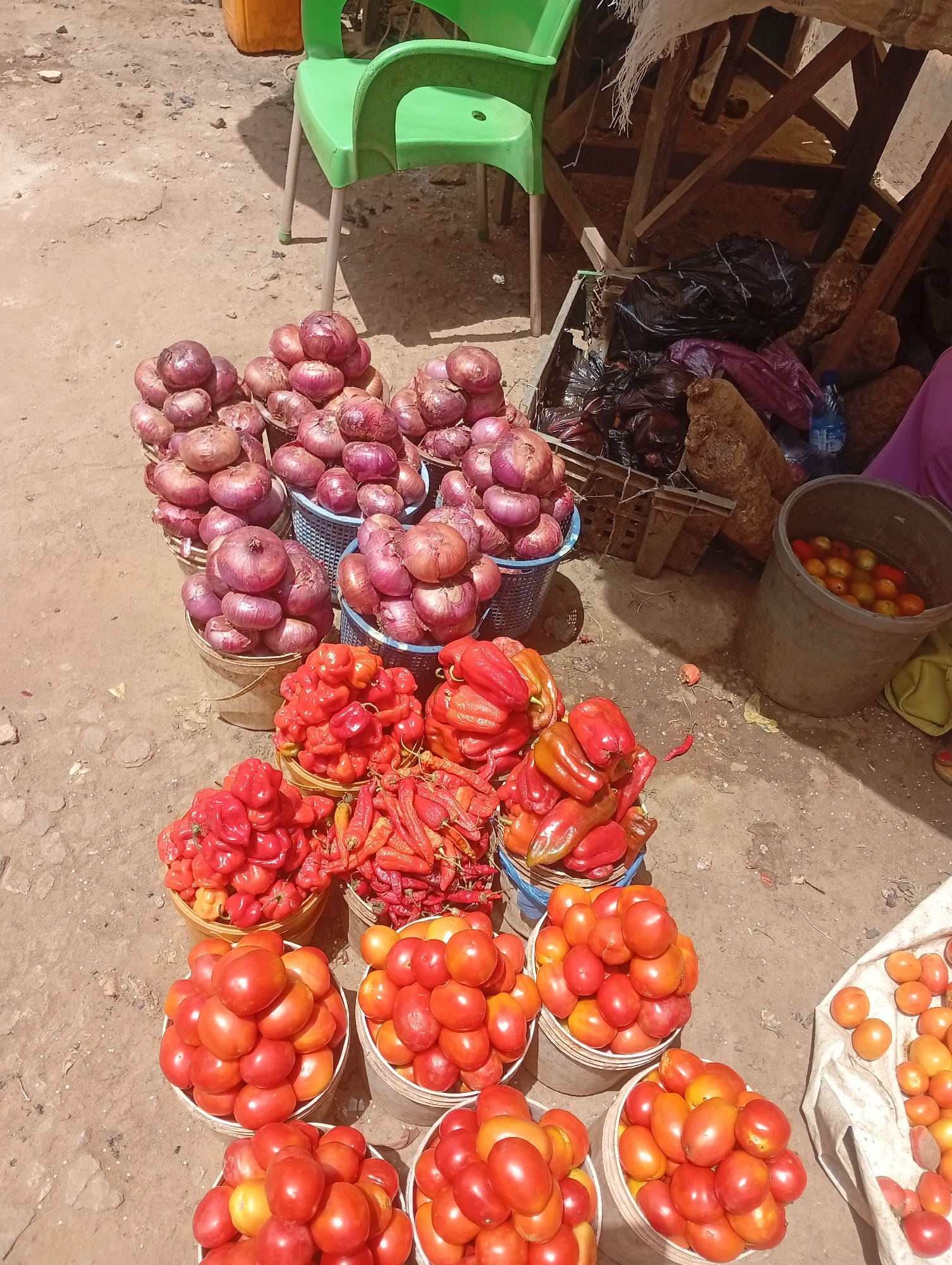 The image depicts a vegetable stall in Barnawa, Nigeria. Piles of red onions are arranged in blue and white plastic buckets. Below the onions are several buckets containing various types of peppers and red tomatoes. A small bucket on the right contains smaller tomatoes submerged in water. The vegetables are arranged on the ground, near a wooden stall structure. A green plastic chair sits in the background. The ground appears to be dusty and made of a compacted, reddish soil.