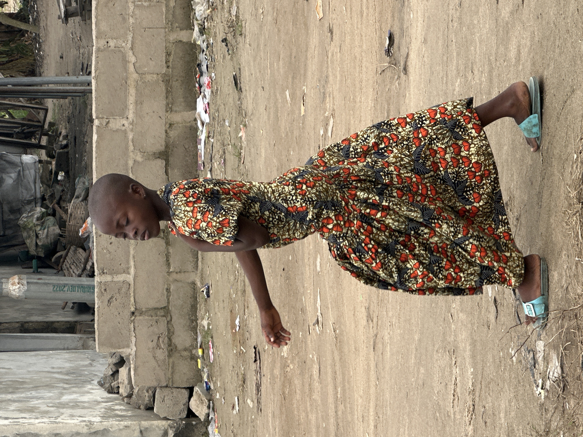 A young Black child, likely a girl, walks from left to right in the foreground of the image, her body angled towards the left. She has a shaved head and is wearing a brightly colored dress with a pattern of red flowers and black and white elements. The dress is full and gathered at the waist, flaring out towards the hem. She is wearing blue slide sandals.
The child is walking on a dirt path that appears to be in an urban or semi-urban environment. Behind her, a low wall made of concrete blocks runs horizontally across the frame. Beyond the wall, there is debris, including plastic bags and scattered papers. Further in the background, there are structures that appear to be buildings or unfinished constructions. The overall environment suggests a somewhat dilapidated or developing area within the city of Badagry, Nigeria. The lighting suggests it is daytime.
