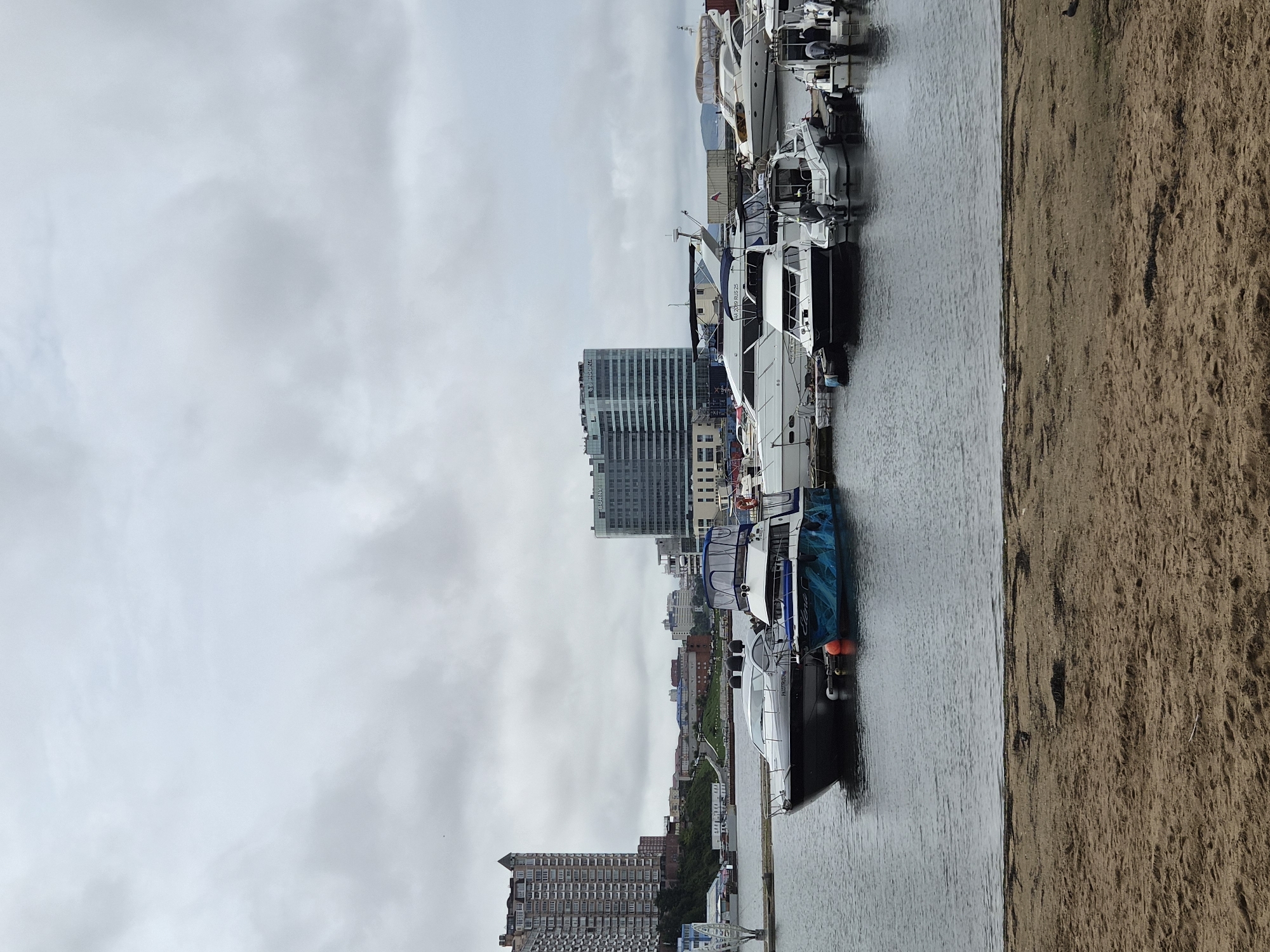 The image depicts a cloudy day at a marina in Vladivostok, Russia. In the foreground, a sandy beach transitions into the calm, gray water of the bay. Several yachts and smaller motorboats are moored side-by-side at docks. One of the boats in the foreground has a distinctive blue and teal wave pattern on its hull and the name "Elena" visible. Further in the background, the city skyline is visible with several tall, modern buildings, including a prominent, light-colored high-rise on the left with many windows and balconies, and a darker, glass-fronted building to its right. The overall scene conveys a sense of a typical urban waterfront on an overcast day.
