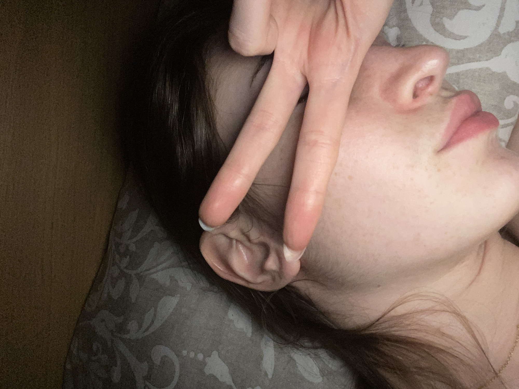 A close-up, high-angle shot depicts a person lying on a patterned pillow. The person's head is turned to the side, revealing their nose, lips, and ear. Two fingers are positioned near the ear, forming a V-shape. The person has dark hair and fair skin with freckles. A portion of a wooden surface is visible in the upper-left corner. A gold necklace is partially visible near the lower right of the frame.