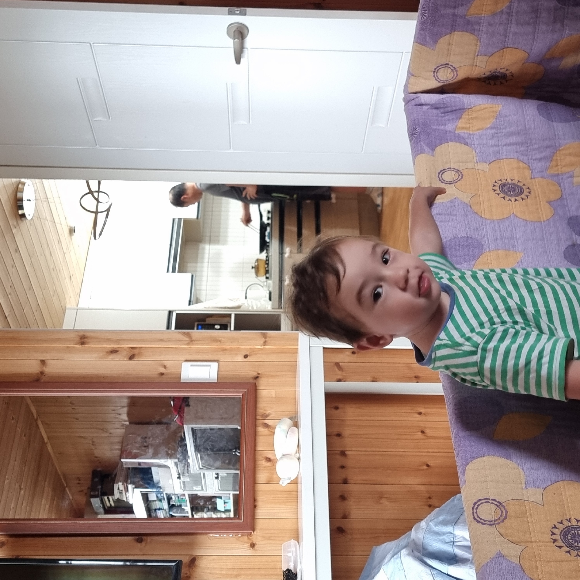 A young child with brown hair and dark eyes, wearing a green and white striped t-shirt, sits in the foreground, leaning on a purple blanket with yellow floral patterns. The child is positioned in front of a wooden wall. Behind the child, a white shelf is visible. To the left of the shelf, a mirror reflects a cluttered room with shelves filled with various items, including boxes and what appear to be books. Further in the background, a person is visible in a kitchen area, appearing to be cooking. A white door with a silver handle is to the right of the frame. The environment suggests an indoor residential setting, possibly in Dunnae-myeon, South Korea, given the distinct wooden paneling commonly found in Korean homes.