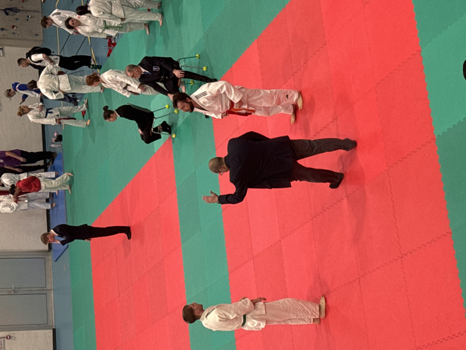 This image depicts an indoor sports facility in Brumath, France, featuring a martial arts competition or training area. The floor is covered with interlocking foam mats, primarily red in the central foreground and midground, surrounded by green mats.
In the immediate foreground, on the red mats, three individuals are central. A male in a dark suit jacket and grey trousers stands with his back to the viewer, his right arm raised with an open palm, seemingly giving a signal. To his left, a male judoka in a white judogi with a green belt stands barefoot, facing right. To his right, a male judoka in a white judogi with an orange/red belt stands barefoot, facing left; an adidas logo is visible on his left pant leg.
In the midground, on the green mats, several individuals are present. To the far left, a male in a dark suit stands with his right hand near his face/ear. Two individuals are seated on light-colored chairs: a female in dark attire on the left, and a male in a dark suit with a lapel badge on the right, whose chair legs are fitted with yellow tennis balls.
Further in the background, numerous individuals are scattered across the green mats. Most are wearing white judogi with various colored belts (including blue, green, and white). Some individuals wear casual attire, such as a person in a red hooded sweatshirt bowing to another judoka, a person in a purple shirt, and an individual wearing a grey baseball cap. A person with a backpack is also visible.
The background includes a light-colored wall or door structure on the left and a partial view of a climbing wall on the right. Small items, possibly water bottles, are visible on the green mat in the background.