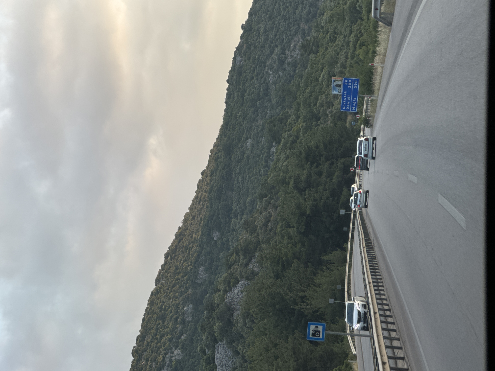 The image displays a multi-lane highway passing through a mountainous, heavily vegetated region under an overcast sky. The scene is located in the Döşemealtı district of Antalya, Turkey, as indicated by a prominent road sign directing towards Korkuteli (36 km), Denizli (200 km), and Muğla (290 km).
From the perspective of a vehicle, possibly the front passenger seat, the bottom edge of the image shows a portion of the vehicle's interior. The asphalt highway occupies the midground, featuring painted lane markings. Metal guardrails line the left side of the main highway, which runs parallel to an elevated road section or ramp, separated by a concrete barrier topped with an additional guardrail.
Several vehicles are observed on the road. On the elevated ramp to the left, a white Peugeot 308 is visible, with a license plate partially obscured but beginning with "07". Further down the main highway, a silver sedan is in the right lane, followed by a dark-colored vehicle and other indeterminate vehicles in the distance.
Two blue traffic signs are prominent. On the left, a square blue sign with a white border displays a white camera icon, indicating a traffic or speed camera zone. On the right, a rectangular blue directional sign provides the aforementioned distances to Korkuteli, Denizli, and Muğla. Above this sign, a smaller, indistinct billboard with a multi-colored image is partially visible.
The background is dominated by steep mountains covered in dense green foliage and scattered rock formations. The sky is largely overcast with grey clouds, showing a subtle yellowish hue towards the upper right, suggestive of either sunrise or sunset light.
