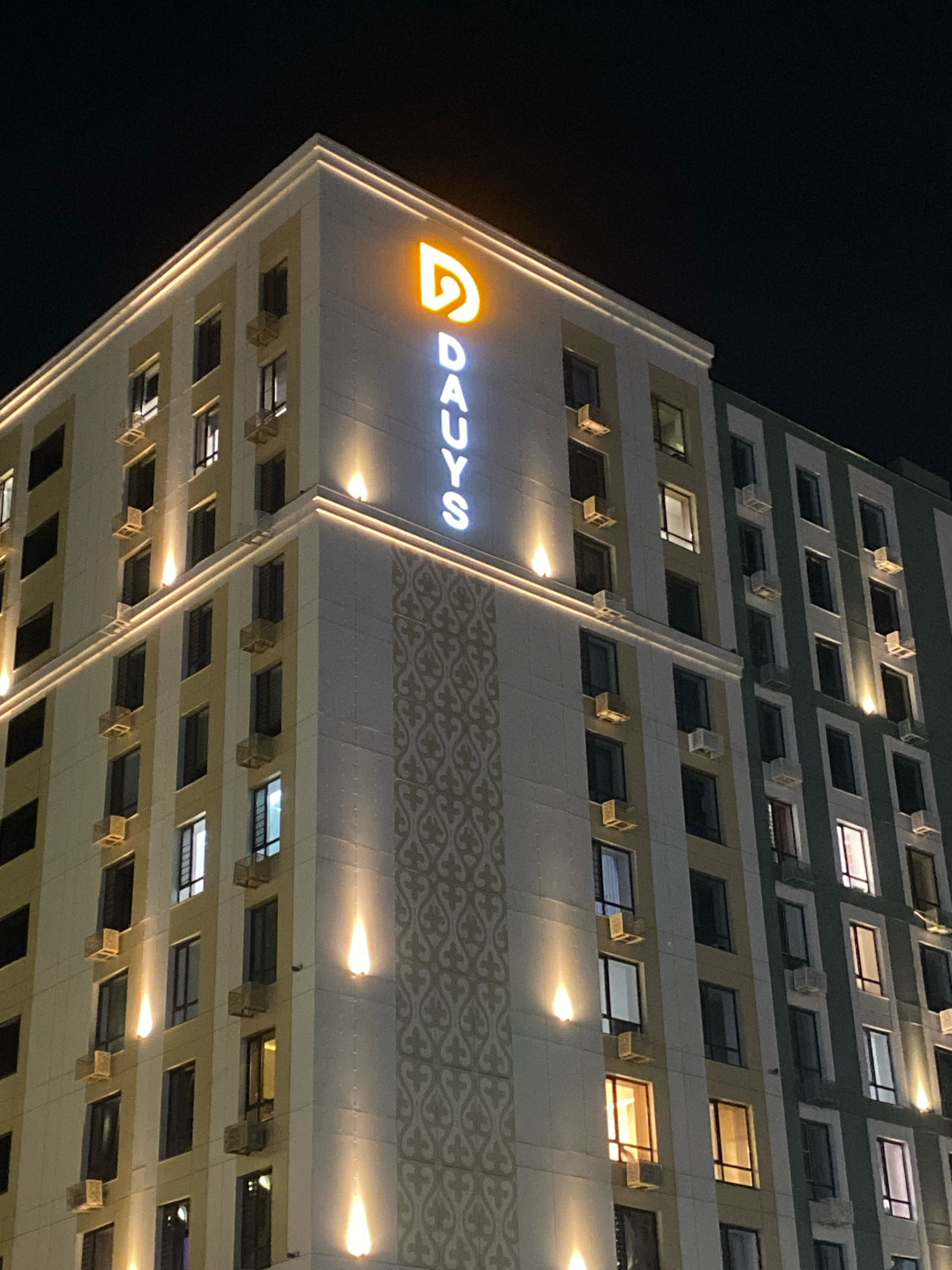 The image shows the exterior of a multi-story building at night, likely a hotel, in Astana, Kazakhstan. The building features a modern architectural design with a light-colored facade accented by golden trim and vertical lighting. A large, illuminated sign on the top facade spells out "DAUYS" in white lettering, with a prominent orange logo above it. Numerous windows are visible, some lit from within, indicating occupancy. Several windows have air conditioning units installed. Decorative patterned panels are integrated into the facade. The overall impression is one of a well-maintained commercial property presented at night.