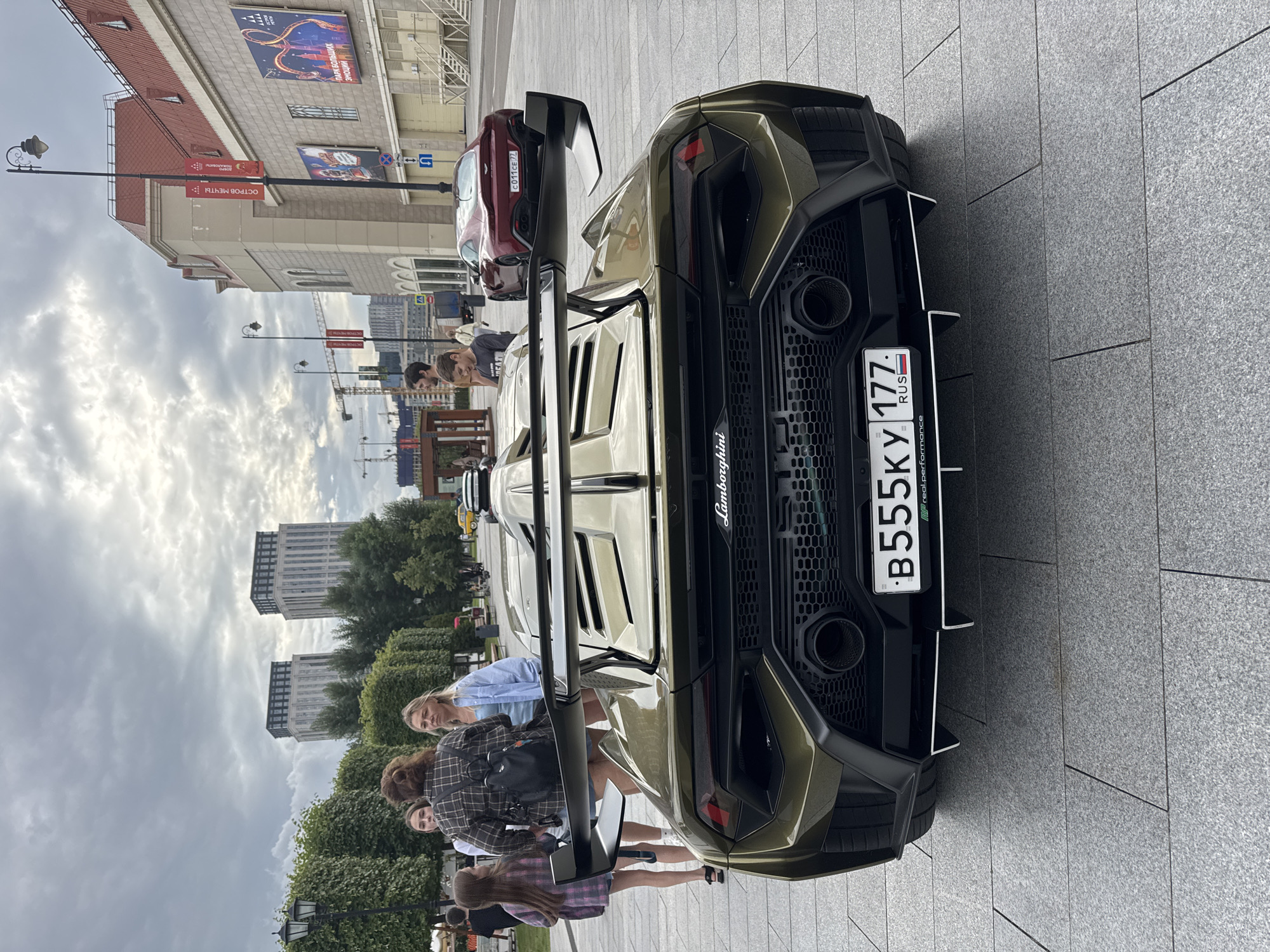 A group of young people are gathered around the back of a green Lamborghini parked on a stone-tiled plaza in front of a building. The car has a prominent rear spoiler and an exposed engine cover with intricate detailing. A license plate with Russian characters and numbers "B555KY 177" is visible, along with the text "realperformance" and "Lamborghini" on the car's rear. In the background, other vehicles, including a red Aston Martin, are visible, as well as more of the building complex and trees. In the distance, modern high-rise buildings stand against a cloudy sky. The weather appears overcast, and the time of day is likely daytime. The people, seemingly in their late teens or early twenties, are casually dressed and appear interested in the car, with some leaning in to get a closer look. The scene suggests a public area, possibly a street or a plaza, in Moscow, Russia.