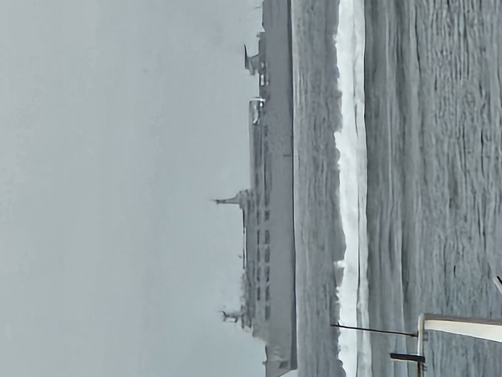 A ferry ship is at sea under a cloudy sky. The ferry is a large, elongated vessel with multiple decks and a tall superstructure towards the stern. Waves are visible in the foreground. The image is taken from a low vantage point, possibly from another boat, as parts of a white railing and a dark pole are visible in the bottom left corner. There are no people visible in the image. The scene appears to be in an open body of water. Without specific landmarks or context, pinpointing the exact location in Denpasar, Indonesia is not possible based solely on this image.