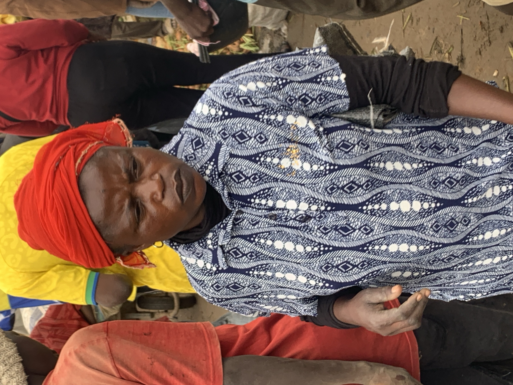 This media file, taken in Gura Top, Nigeria, captures a scene of people gathered, possibly in a market or public space. The primary focus is on several individuals, seen from a top-down perspective.
One person in the foreground wears a red headscarf and a yellow long-sleeved shirt. Their face is visible, and they appear to be looking upwards. To their right, another person is dressed in a blue and white patterned shirt with a black long-sleeved undershirt. Their right hand is visible, resting on their lap. Further in the background, another individual is seen from the waist down, wearing a red top and black leggings. Other individuals are partially visible, suggesting a gathering of people.
The ground appears to be dirt or sand, with some straw-like material scattered around. The overall lighting suggests it might be daytime. The image has a slightly tilted orientation. No visible text is present in the media file. The overall mood of the scene is difficult to ascertain due to the limited facial expressions and body language captured, but it appears to be a candid moment within a community setting.