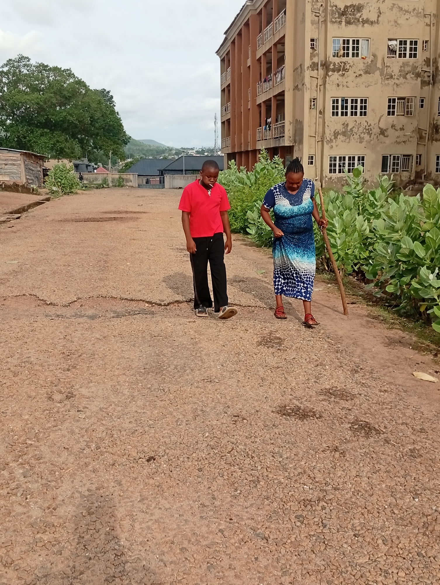 Adankolo, Nigeria: A woman in a blue dress and red sandals walks with a wooden stick along a cracked, gravel road. A young man in a red shirt and black pants walks alongside her. A building with multiple windows and balconies is on the right. Green bushes line the right side of the road. In the distance, there are trees, buildings, and hills under a cloudy sky.