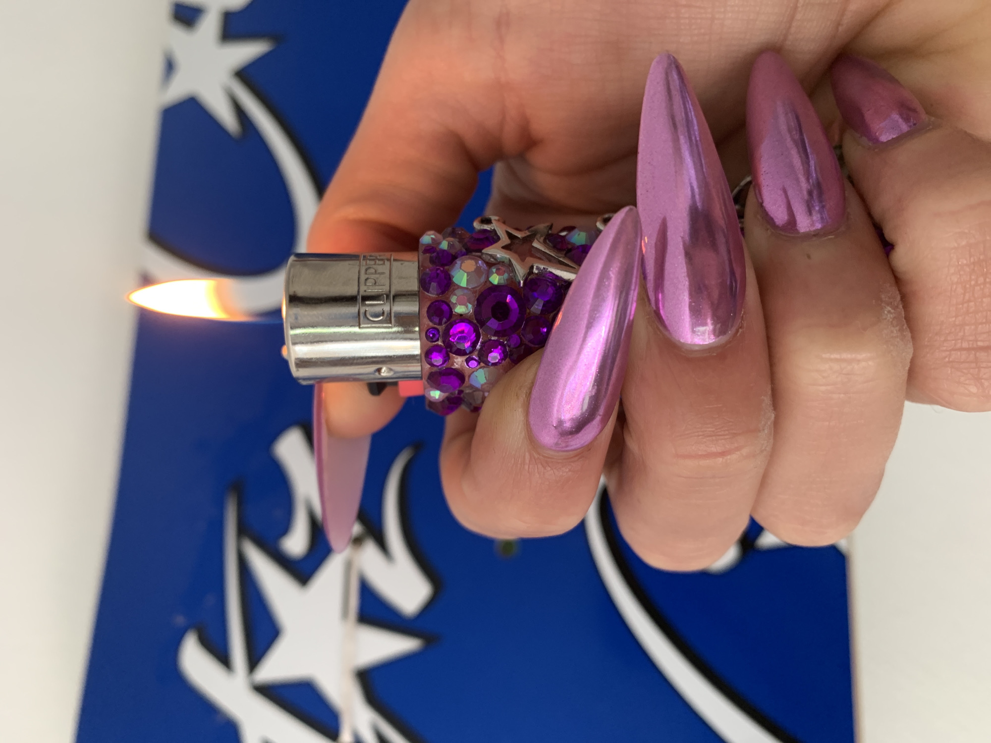 A close-up image shows a hand, likely female, holding and igniting a lighter. The hand features long, pointed, stiletto-shaped fingernails, which are painted in a metallic pink-purple finish that reflects light. The lighter itself is a silver "CLIPPER" brand, heavily embellished with various shades of purple and iridescent rhinestones, and adorned with a small silver star charm attached to its side. A bright orange and yellow flame is actively burning from the lighter's tip. In the background, a distinct blue surface is visible, decorated with multiple white and black outlined star-like shapes, suggesting a logo or a patterned design element. The image captures the moment of the lighter being used, emphasizing the intricate details of the hand's styling and the decorated lighter. There are no discernible cues for the time of day or emotions. The specific location within London, United Kingdom, is not visually indicated by the background elements.