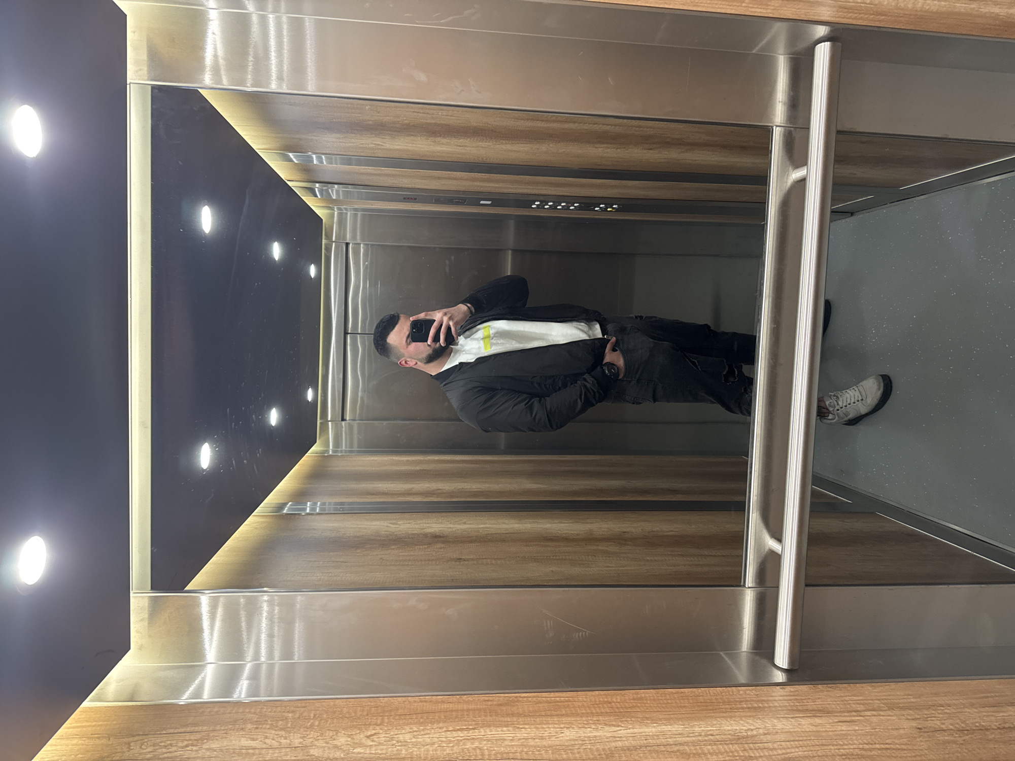 The image captures a man taking a selfie in an elevator. The elevator has wood-paneled walls with recessed lighting on one side, and a reflective stainless steel surface with a silver grab bar on the other. The man is wearing a black jacket over a white shirt, with black pants and grey sneakers. He is holding a black smartphone. The floor appears to be grey. The image does not provide information to identify the location in Sofia, Bulgaria.