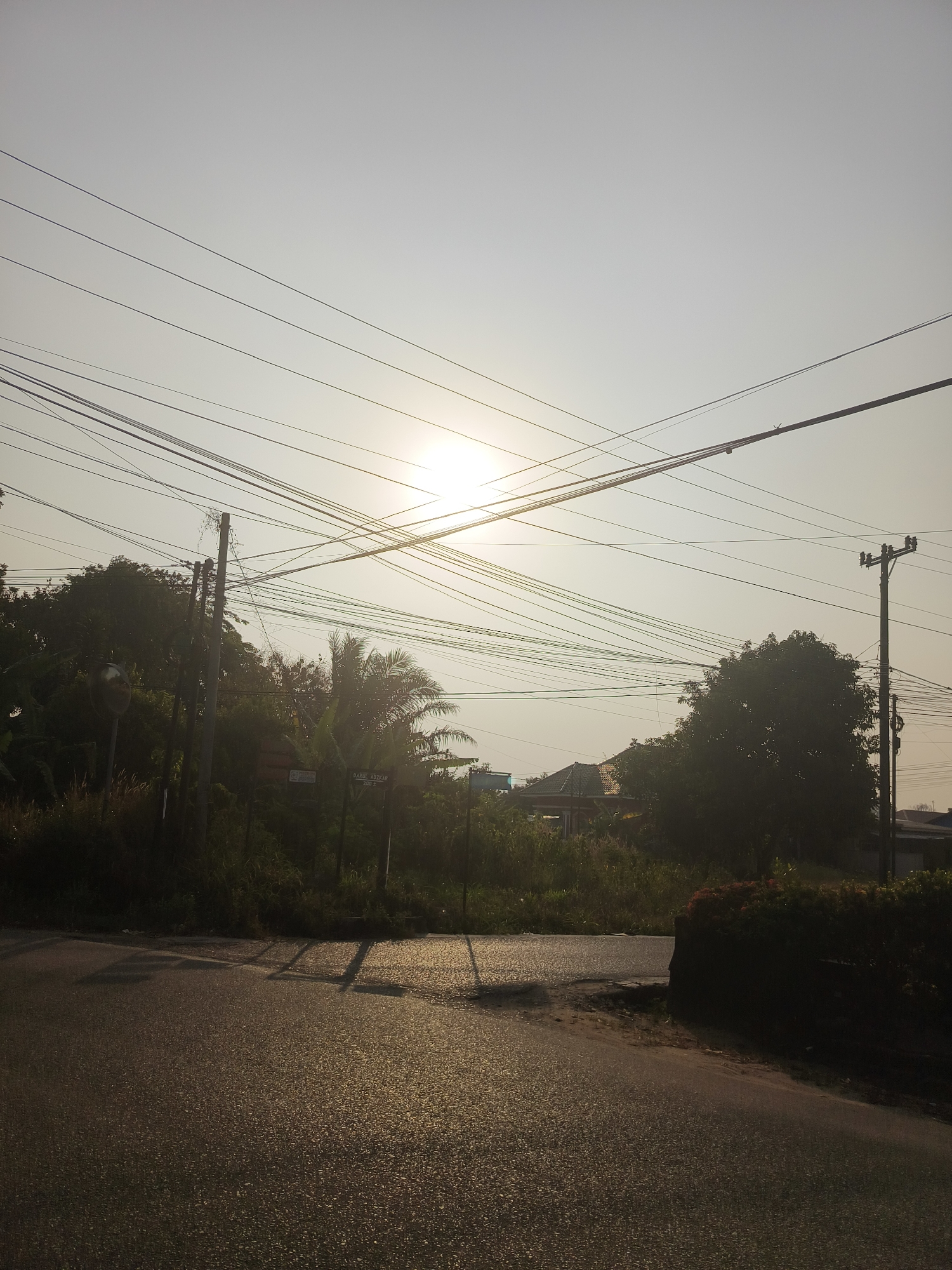 The image depicts a street scene in Pangkalpinang, Indonesia, during what appears to be sunrise or sunset due to the low angle of the sun and its soft, diffused light. The sky is a pale, hazy gray with the sun positioned centrally, casting a gentle glow and creating lens flare. Numerous electrical wires crisscross the upper portion of the frame, supported by utility poles visible on the left and right sides of the image.
The foreground is dominated by an asphalt road, reflecting the sunlight and appearing wet or glistening. Shadows from the electrical wires and possibly nearby foliage are cast across the road surface. To the left and right of the road, lush green vegetation, including trees and shrubs, lines the roadside. The vegetation appears dense and somewhat unkempt in places.
In the midground, behind the vegetation, hints of buildings and structures can be discerned. One building features a tiled roof, and another appears to be a residential house. A street sign is partially visible on the right side of the image, reading "DARUL AZHAR 100 M". A round, reflective traffic mirror is mounted on a pole on the left side of the road, near the vegetation. The overall impression is a somewhat rural or suburban street setting within the city, characterized by a high density of overhead electrical infrastructure and abundant natural greenery.