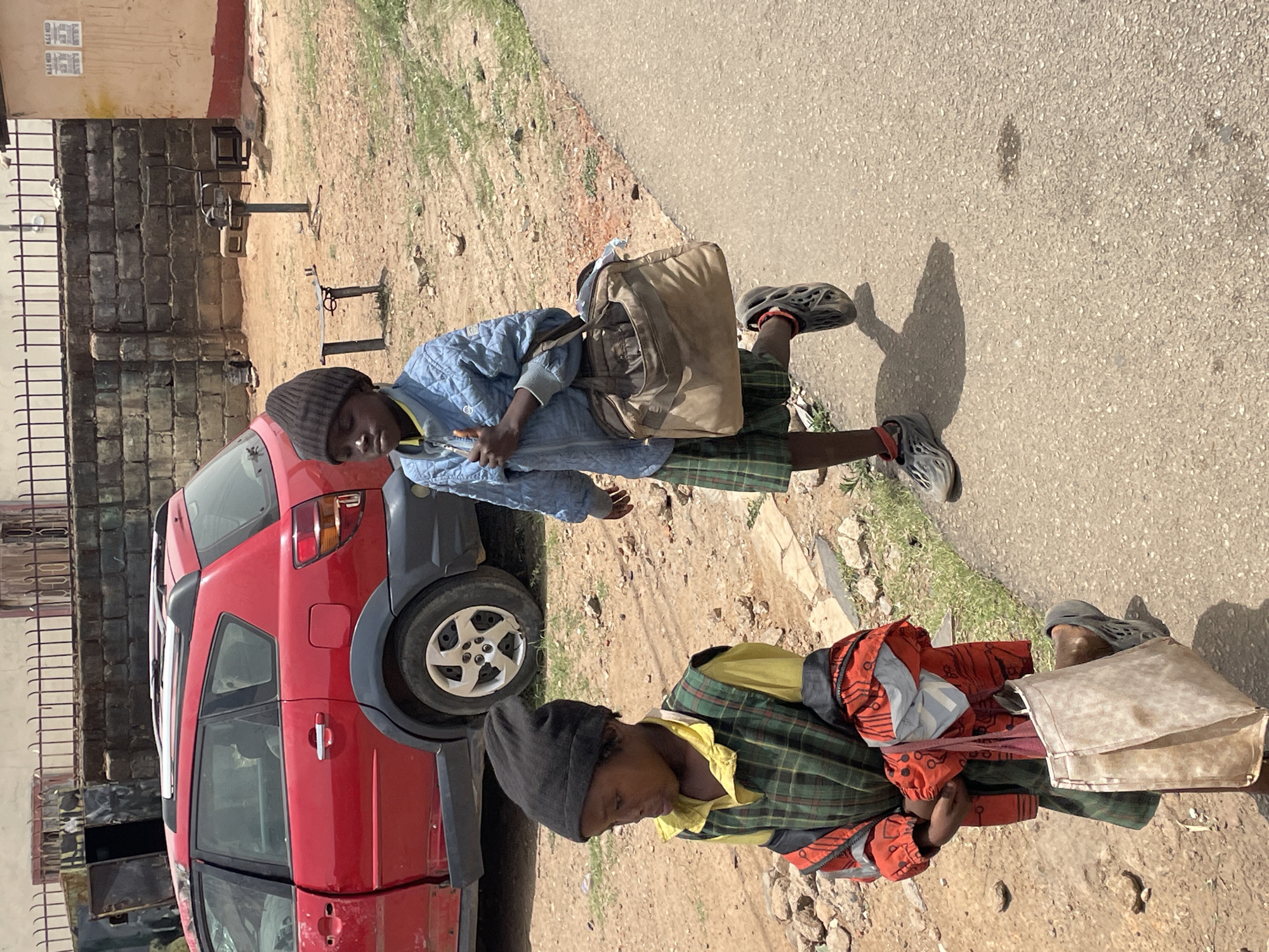 Two people are walking on a street in Jos, Nigeria. One person wears a blue jacket and carries a brown bag. The other person wears a black hat, green plaid shirt, and red jacket, and carries a white bag. A red car is parked nearby. A fence and brick wall are behind the car. The ground is a mix of dirt, grass, and concrete.