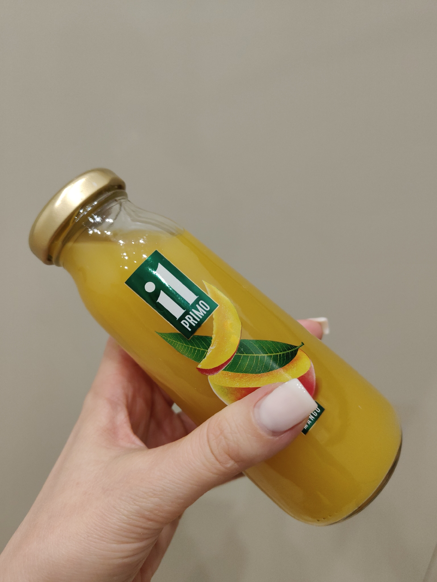 A hand holds a glass bottle filled with yellow liquid, likely juice, against a neutral gray background in an unknown city. The bottle has a gold-colored cap and a label with the text "II PRIMO" and an image of a mango. The hand has fair skin and neatly manicured nails. The scene is well-lit, suggesting an indoor environment.