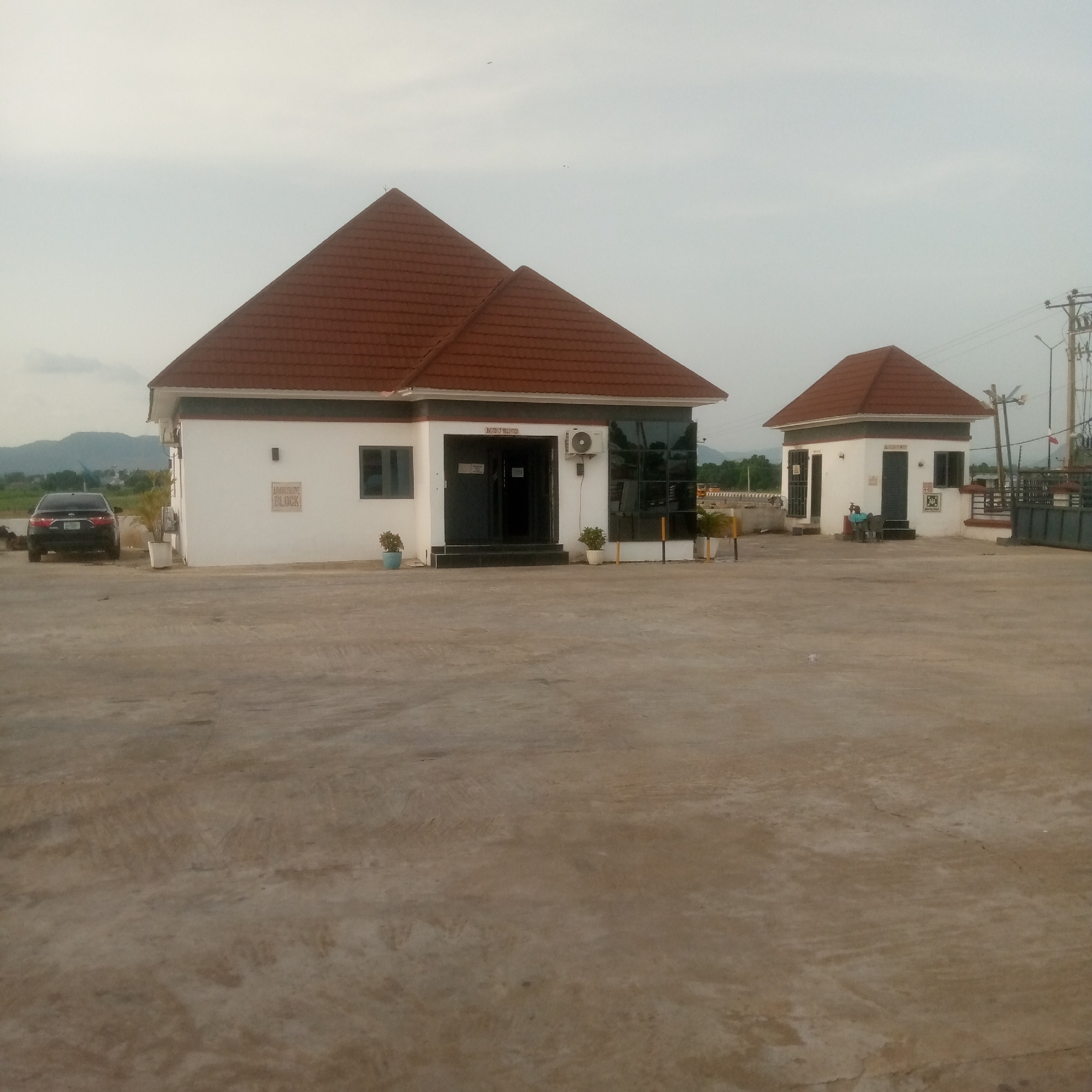 In the city of Unknown, Unknown, the scene depicts two white buildings with terracotta tiled roofs, situated in an outdoor paved area under an overcast sky. The larger building, positioned on the left, features a dark-tiled entrance with a sign indicating "RECEPTION". It has a dark-tinted glass facade on its right side and a dark-framed window on its left. An air conditioning unit is visible above the entrance. To the left of this building, a dark-colored sedan is parked on the pavement. In front of the building, there are potted plants. A smaller, square-shaped building with a similar terracotta roof is located to the right of the larger structure. This building has windows and a doorway, and a person is seated outside it on a bench. The paved area extends to the foreground and appears to be a parking lot or courtyard. In the background, a light pole with electrical wires and a street light can be seen, along with distant hills and trees.