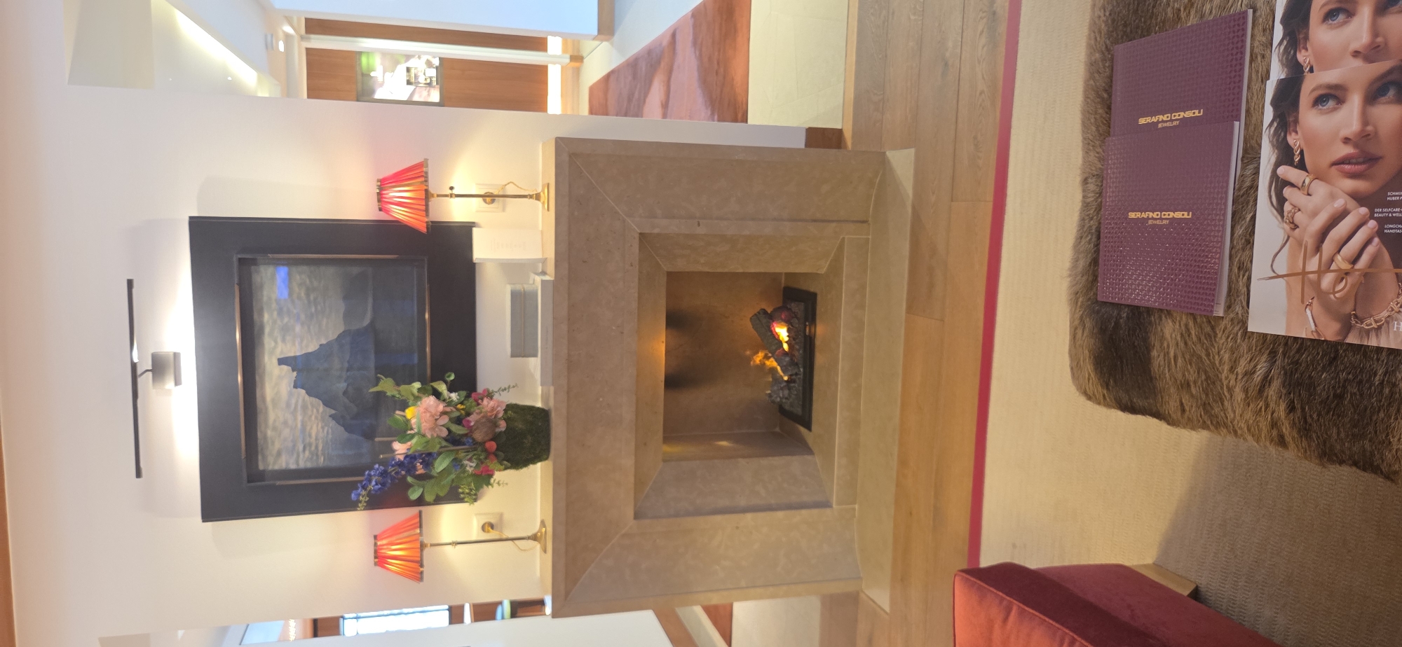 The image depicts an indoor scene featuring a modern fireplace as the central element. The fireplace has a light-colored stone surround and a dark, rectangular frame above it, within which a framed artwork of mountains is displayed. Two red, pleated lamps with gold bases are positioned on either side of the artwork, casting a warm glow. In front of the fireplace, on the mantelpiece, sits a floral arrangement in a moss-covered pot, flanked by two minimalist grey boxes. The fireplace itself contains a lit gas fire with logs.
The flooring is light-colored wood. In the foreground, a plush, brown rug covers part of the floor, with a red upholstered ottoman or chair visible to the left. On the rug, there are two purple booklets with embossed text and a fashion magazine featuring a close-up of a woman's face and hands adorned with jewelry.
The overall ambiance is sophisticated and luxurious, suggesting a high-end interior, possibly a hotel lobby, lounge, or a private residence in a place like Lech, Austria, known for its upscale ski resorts and elegant accommodations. The lighting is warm and inviting, with the fire and lamps creating a cozy atmosphere.