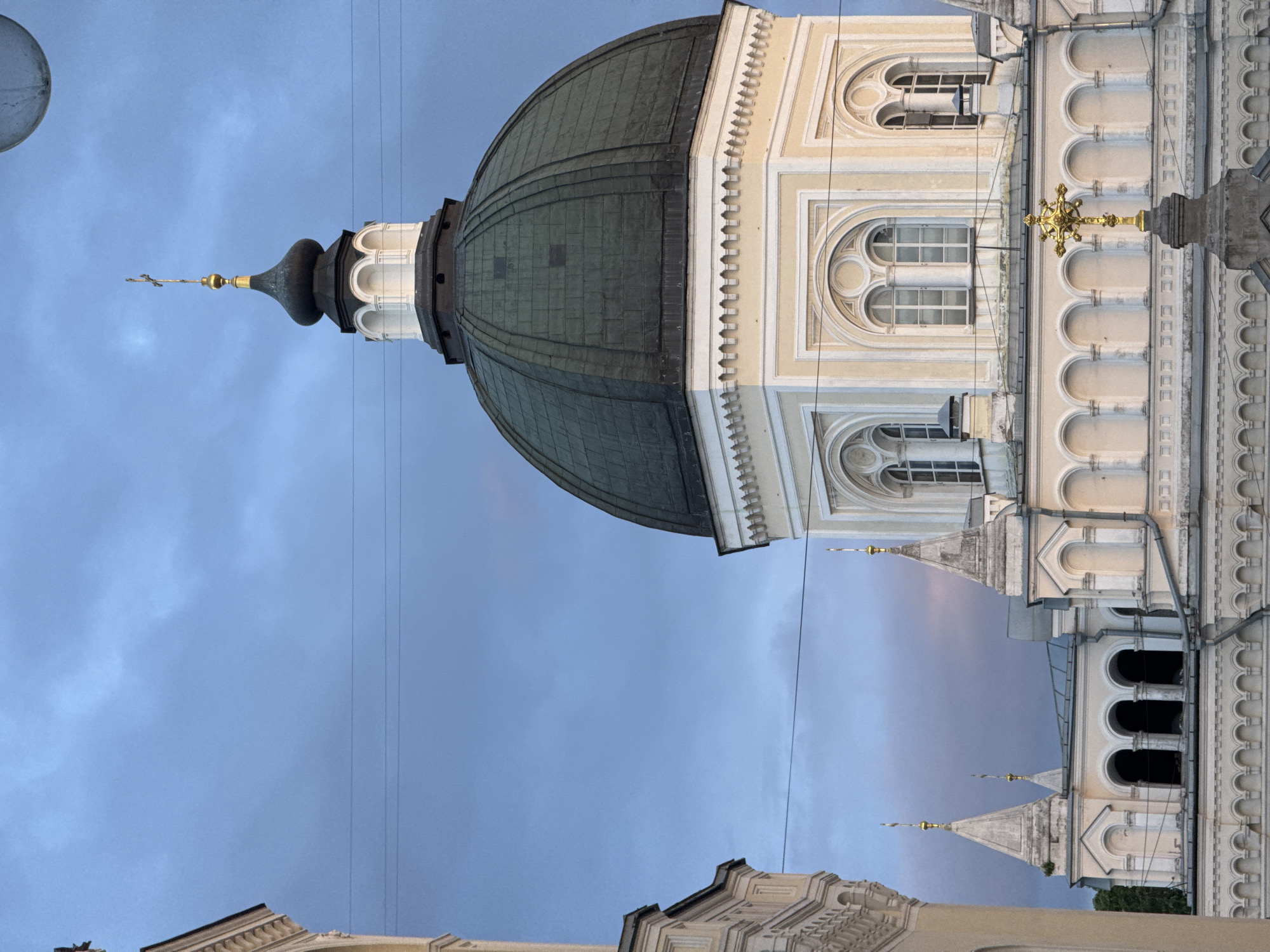 The image shows a portion of the Cathedral of Christ the Savior in Moscow, Russia, taken from a low-angle perspective. The dome, covered in what appears to be copper, is prominent, with its ornate spire topped by a cross extending into the blue sky. The architecture features detailed classical ornamentation, including arches, windows, and columns. Electrical wires are visible against the sky. Another, smaller spire is visible in the distance, as are other parts of the building. The image shows late day and/or early dusk.