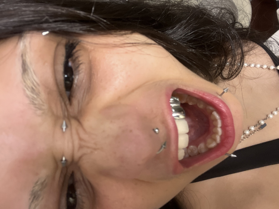 A close-up, angled shot features a person with dark hair and facial piercings. Piercings are visible above the eyebrow, between the eyes, on the cheek, and around the mouth. The person's mouth is open, revealing white teeth, a prominent silver tooth cap, and a red tongue. A pearl necklace is partially visible on the person's neck, along with a black strap. The overall image is unconventional, focusing on facial modifications.