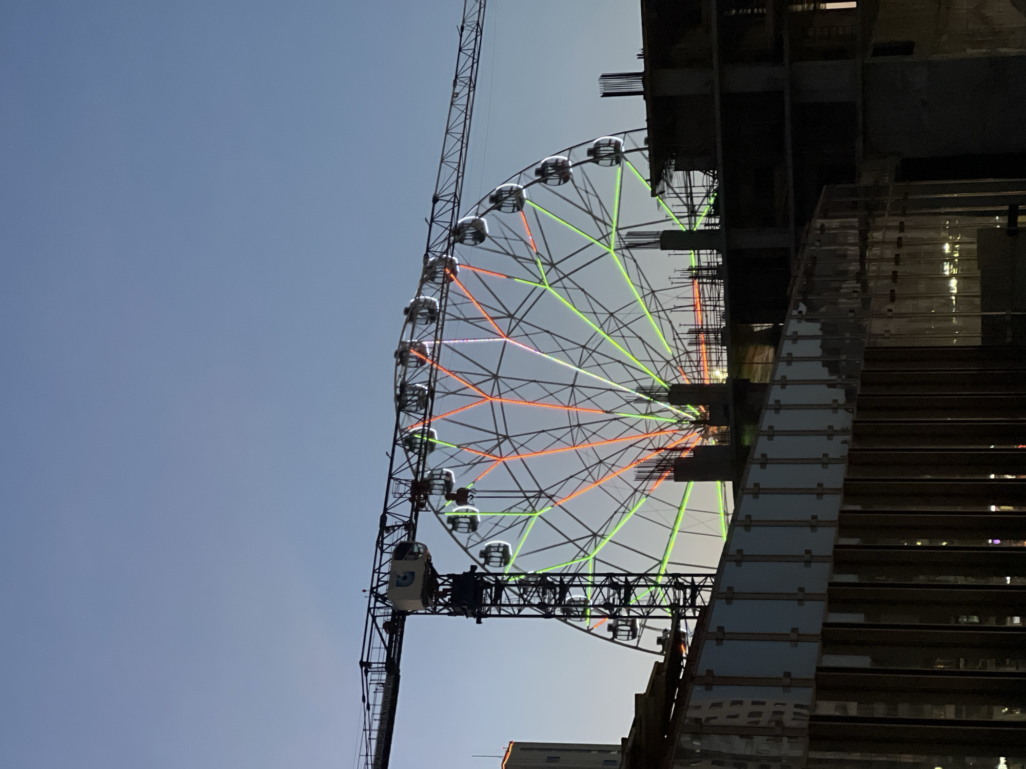 This vertically oriented image depicts a large Ferris wheel standing alongside a modern building under construction, set against a clear, bright blue sky.
The Ferris wheel, partially visible, dominates the central and left areas of the frame. Its intricate metal framework is illuminated with multi-colored lights, predominantly orange/red and green, highlighting its spokes and structural elements. Numerous enclosed gondolas (cabins) are suspended from its circumference; one white gondola on the left displays a faint, illegible blue symbol or number. A robust metallic support structure, possibly a crane or part of the Ferris wheel's permanent base, is visible on the far left.
To the right, a multi-story building is actively under construction. Its dark concrete structure and exposed rebar are visible, especially towards the top. A significant section of its facade features dark, reflective glass panels, which reflect some light and show faint interior illumination.
The scene appears to be set during late afternoon or early evening, given the bright sky and the active illumination of the Ferris wheel's lights. The weather is clear with no clouds visible. The image conveys a sense of urban development and modern infrastructure in Kazan, Russia. No people are visible.