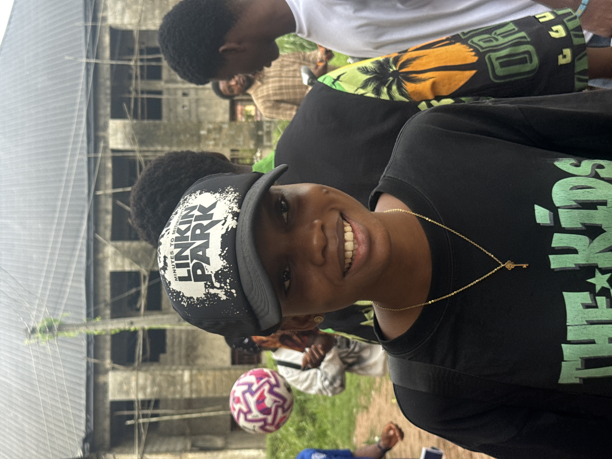 In the city of Ihiagwa, Nigeria, a young Black woman in a black t-shirt and a black baseball cap with "LINKIN PARK" emblazoned on it, stands center frame, smiling. She wears a delicate gold chain with a key pendant. Behind her, other individuals are partially visible, including a person in a white t-shirt on the right and another in a brown plaid shirt further back. A colorful soccer ball hovers in the air to the left. The background features a partially constructed building with visible scaffolding and a grey corrugated metal roof, suggesting ongoing construction. Overhead, electrical wires crisscross the sky, and a utility pole is entwined with green foliage, hinting at an outdoor, possibly urban or semi-urban setting. The lighting is overcast, suggesting a cloudy day.