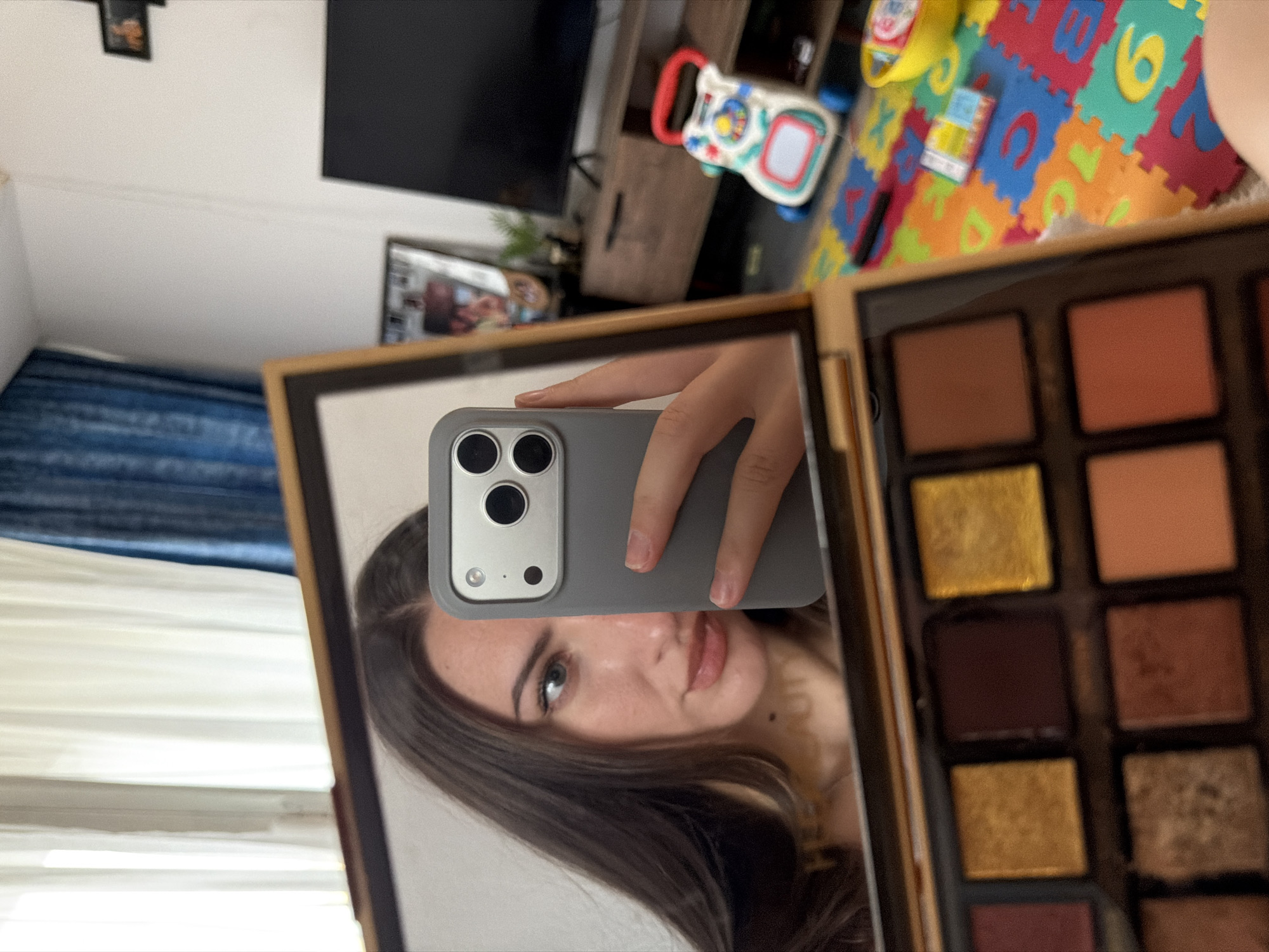 In Sharm El Sheikh, Egypt, a young woman with long, dark hair and light-colored eyes is captured in a mirror reflection, holding a grey smartphone up to take a selfie. She is looking to the side with her lips slightly parted, wearing subtle makeup. In the foreground, a palette of eyeshadows with various shades of brown, gold, and burgundy is visible. The background shows a living room with white and blue curtains, a wooden TV stand with a black television, a child's colorful play mat with letters and numbers, and a red and white push toy. The overall scene suggests a casual moment at home.