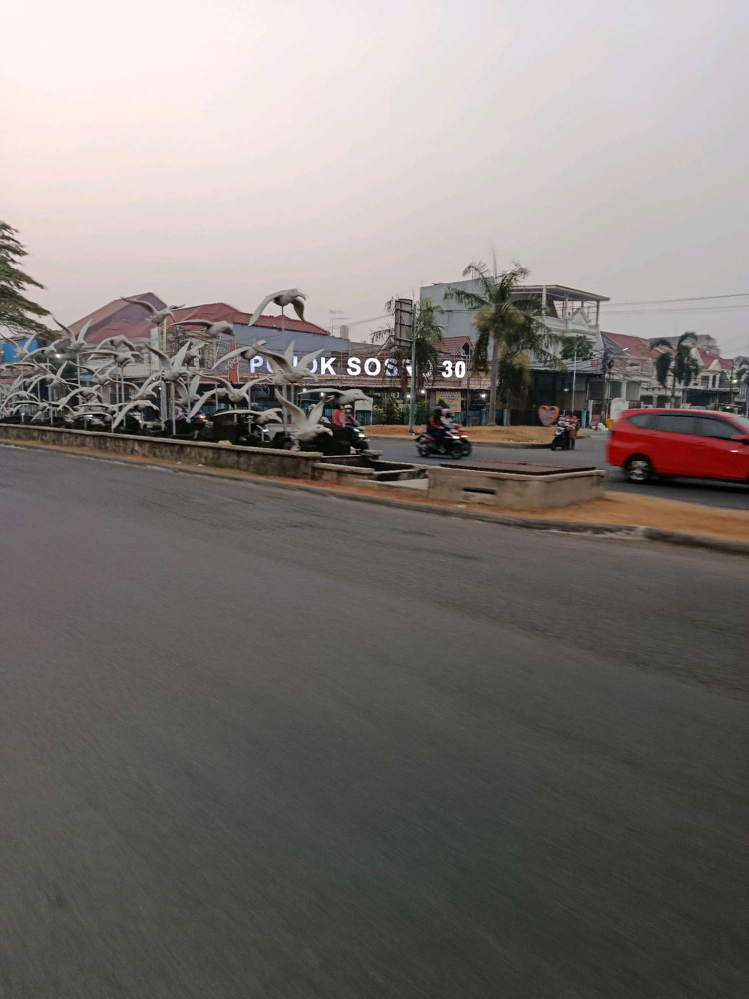 The image captures a street scene in Bekasi, Indonesia, likely in the Rawa Lumbu area, suggested by the prominent "JLUK SOS" sign with the number "30." The foreground is dominated by a blurred asphalt road, indicating motion. In the mid-ground, a distinctive sculpture of numerous white birds in flight is visible along the side of the road. Behind the sculpture, there are buildings with red roofs and modern architectural elements, including a building with multiple stories and balconies, as well as palm trees. The sky is overcast with a pale, hazy light, typical of dawn or dusk. Traffic is present, with a red SUV and several motorcycles visible. One motorcycle carries two individuals, and another motorcycle has a single rider. A heart-shaped decoration is also present on a roadside planter. The overall impression is a contemporary urban environment with a notable public art installation.