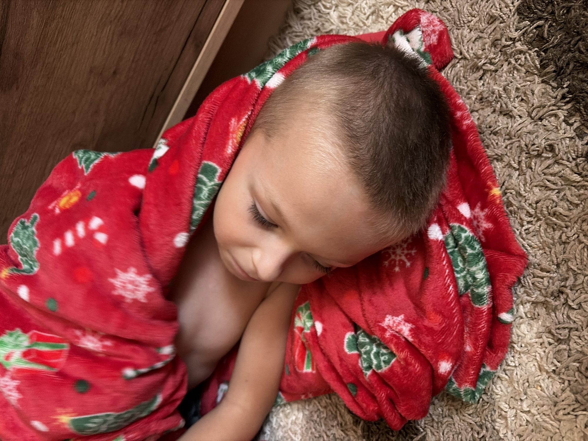 A young child is sleeping, wrapped in a red blanket with a Christmas pattern. The child's head is resting on a beige carpet, and a wooden surface is visible in the background. The scene appears to be indoors.