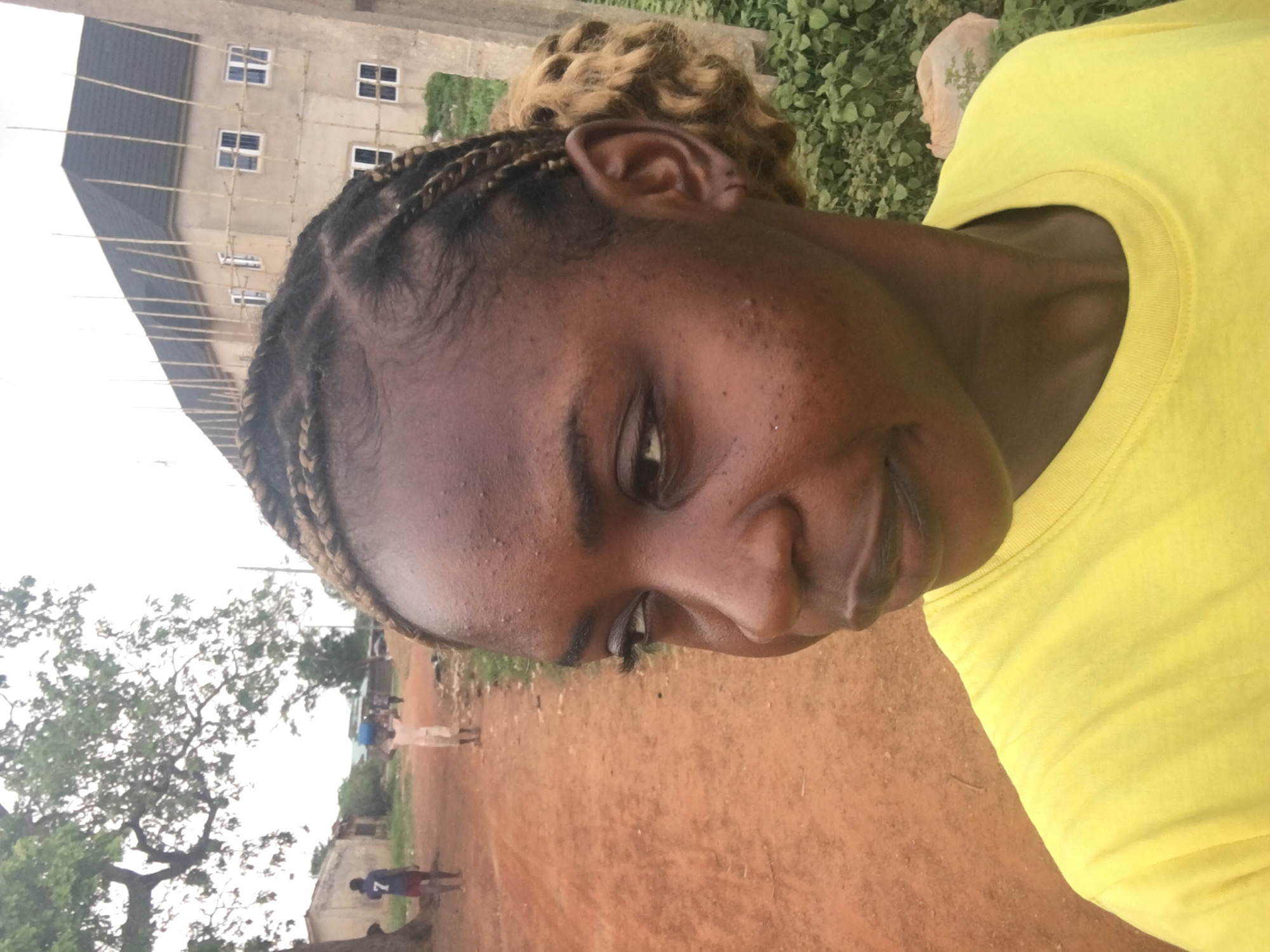The image captures a slightly tilted, close-up outdoor portrait of a young person, likely a woman, with dark skin and a gentle, subtle smile. She is wearing a bright yellow t-shirt. Her hair is styled in cornrows with a bun of lighter, curly hair at the back of her head, just above her right ear.
The setting appears to be a rural or semi-urban area in Enyibichiri, Nigeria. The foreground and mid-ground consist of reddish-brown dirt or earth. In the background, there are various elements: a large tree with sparse foliage on the left, and a multi-story building on the upper left, which appears to be under construction or renovation, featuring visible scaffolding and several rectangular windows. Greenery and bushes are scattered around the environment.
Further in the distance, on a dirt path, several indistinct figures are visible. One figure stands out wearing a blue shirt with the white number "7" clearly printed on the back. The sky is overcast, providing soft, diffused natural light to the scene. The image conveys a calm, everyday moment in an outdoor local setting.