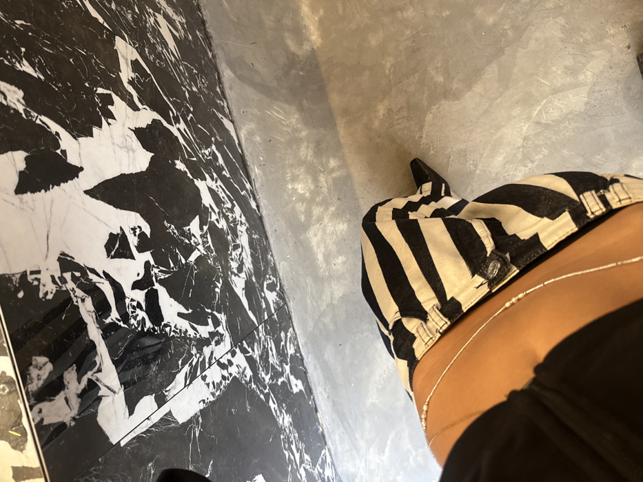 The image depicts the lower body of an individual in an indoor setting. The subject, displaying a dark skin tone, is seated and wears black and white horizontally striped pants. A thin, beaded body chain with small, light-colored accents is visible wrapped around their midriff and lower abdomen, situated above the pants. A portion of a black top is visible on the left, covering the upper abdomen. A black, pointed-toe shoe or boot is partially visible on the subject's right foot. The background features a large, vertical panel of black and white veined marble, exhibiting irregular patterns. The floor is composed of a light grey, uniform concrete or polished cement surface. The environment suggests a contemporary interior space. The specific location within Paris, France, cannot be determined from the visual information provided.