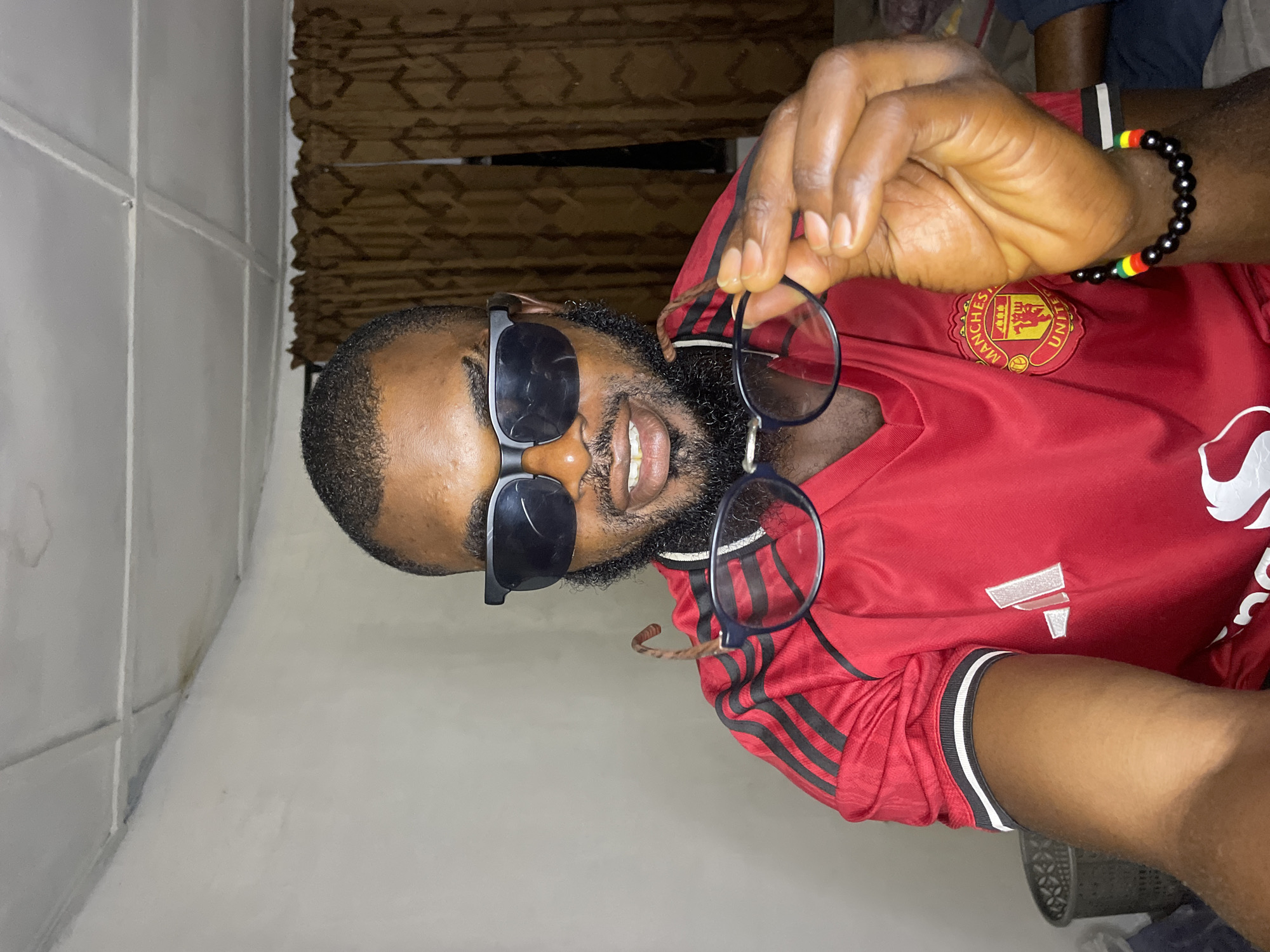 A young Black man with short hair and a beard is indoors, holding up a pair of glasses in his right hand. He is wearing a red Manchester United jersey with black stripes on the sleeves and the team crest on the chest. He is also wearing a pair of black sunglasses. The man is looking at the camera with a slight smile. The background appears to be a room with beige walls and brown curtains. The lighting suggests it is nighttime or indoors. The image does not provide any specific information about the location within the city of Jos, Nigeria.