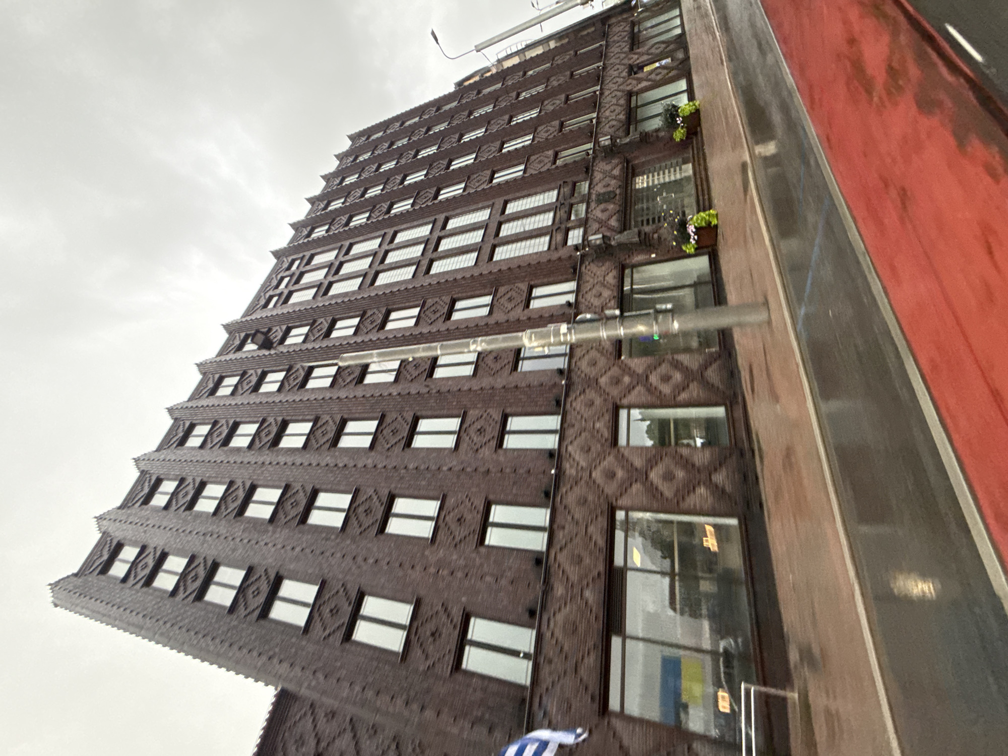 A tall, dark brick building with a stepped, art deco facade dominates the frame, viewed from a low angle on a wet, overcast day. The building features numerous rows of windows with white frames and a decorative diamond pattern etched into the brickwork on several levels. A Ukrainian flag, blue and yellow, is visible on the left side of the building, partially draped and fluttering. The foreground shows a wet red surface, possibly a sidewalk or a lane marker, and a dark, reflective wet surface, likely a road or a flooded pavement. A single lamppost stands on the right side of the frame, partially obscuring the building. The overall impression is of a historic building in an urban setting on a dreary day. This building is located in the city of Tallinn, Estonia.
