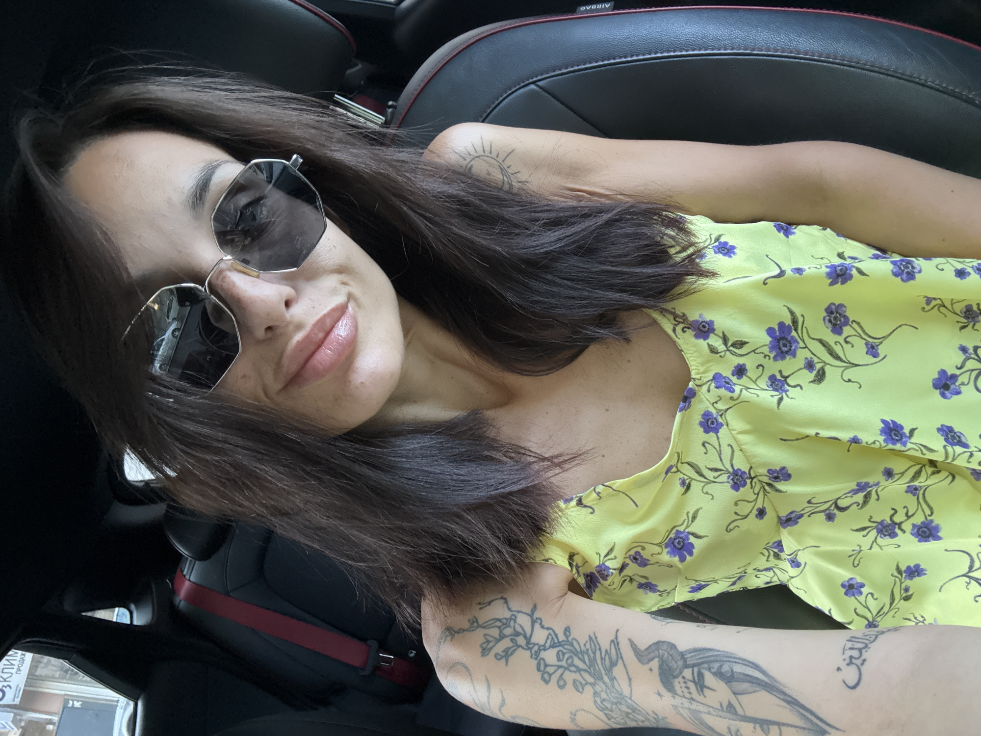 The image shows a woman sitting in the driver's seat of a car. She is wearing large, modern sunglasses and a yellow floral dress. She has long, dark hair and several tattoos on her arms. The car interior is visible, with black leather seats and a red seatbelt. A building with Cyrillic lettering is reflected in her sunglasses, suggesting the location is a city in Bulgaria. The scene appears to be a self-portrait taken inside a vehicle, likely a car, during daylight. The woman is looking towards the camera, with a slight smile.