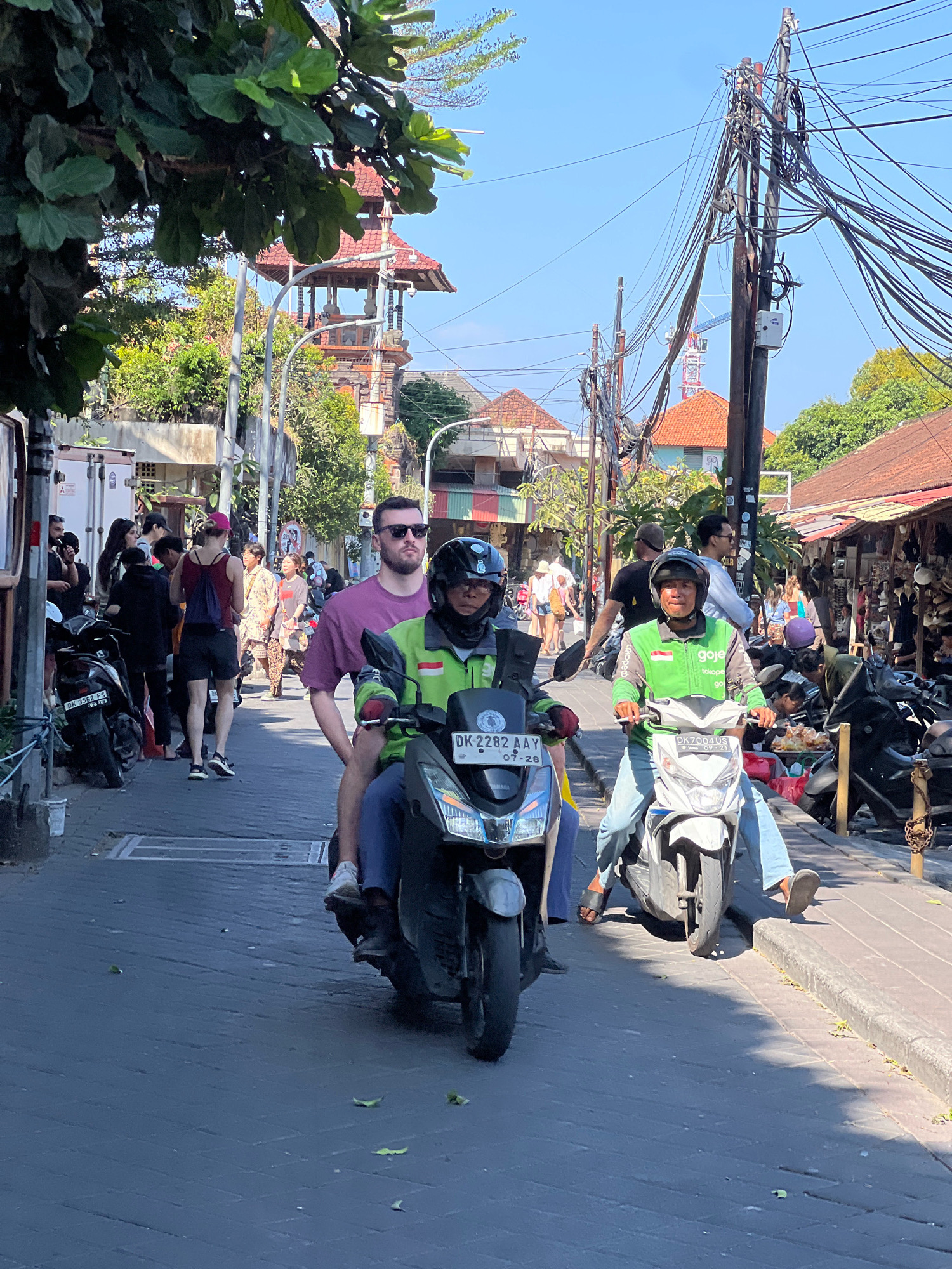 The image captures a street scene in Kuta, Indonesia, on a bright, sunny day. The foreground is dominated by two motorbikes, one in black and one in white. The black motorbike is ridden by a person wearing a green jacket with a "gojek" logo and blue jeans, facing forward with a helmet. Behind them, another individual in a purple t-shirt with sunglasses is seated on the motorbike. The white motorbike is ridden by a person in a green jacket with a "gojek" logo, blue jeans, and a helmet, also facing forward.
To the left of the motorbikes, a man wearing a purple t-shirt and sunglasses stands on the street, looking to his right. Behind him, a woman in a red tank top and black shorts with a blue backpack walks away from the camera. Other pedestrians are visible further down the street, some on foot and others on motorbikes.
The environment is characteristic of a busy urban area. Buildings with red-tiled roofs line the right side of the street, with shops displaying goods. To the left, more buildings and parked motorbikes are visible. Overhead, a dense network of power lines and utility poles crisscrosses the sky. In the background, a traditional Balinese-style tower with a red roof stands out against the clear blue sky, and lush green trees provide some foliage. The street itself is paved with dark bricks. The overall impression is one of a vibrant, bustling street in a tropical location.