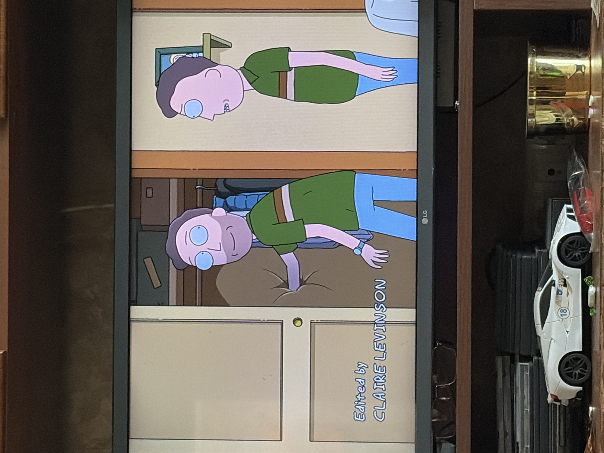 An animated television screen displays a scene from "Rick and Morty." Two characters, resembling Jerry Smith, are depicted in a room. The character on the left, facing right, wears a green shirt with brown and white stripes and blue pants. He is reaching into a large, tan-colored object. The character on the right, facing left, is also wearing a green shirt with similar stripes and blue pants. Text on the lower left of the screen reads "Edited by CLAIRE LEVINSON." The background includes a closet with clothes and a small framed picture on a shelf. Below the television, a shelf holds a white toy sports car with the number "18" and "AEW" logo on its side, along with a gold cylindrical container. The television is placed on a wooden shelf unit. The scene appears to be indoors.