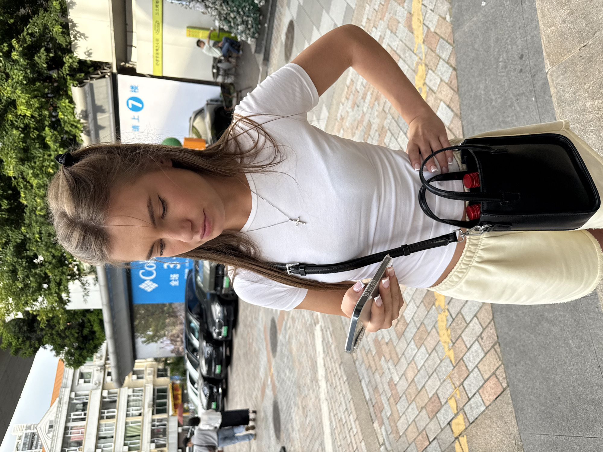 A young Caucasian woman is standing on a paved sidewalk in what appears to be an urban setting in Haizhu District, China. She is wearing a white t-shirt and light-colored shorts, and she is holding a silver smartphone in her right hand, looking down at it. A black crossbody bag with two red-capped bottles inside is hanging from her left shoulder. In the background, several cars are parked in a line, and a building with multiple windows is visible. Trees and an overhead structure also contribute to the urban landscape. The lighting suggests it is daytime.