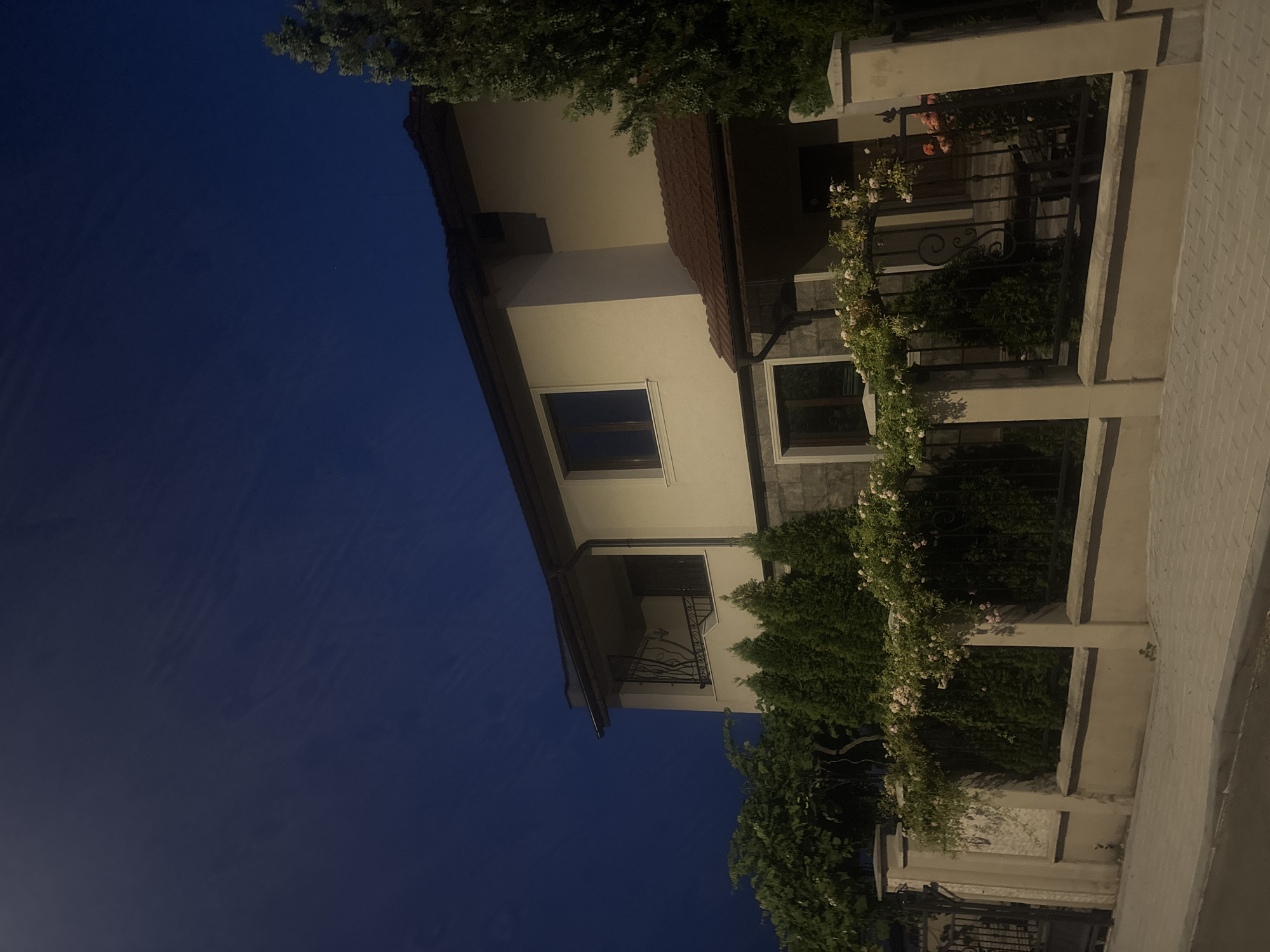 The image depicts the exterior of a two-story house at dusk, located in Burgas, Bulgaria. The house is constructed with a light-colored facade and a brown tiled roof. It features a balcony on the upper floor with a decorative wrought-iron railing, and multiple windows on both levels. A covered porch area is visible near the entrance on the right side of the house. The property is enclosed by a fence, a portion of which is made of stone pillars and a wrought-iron railing adorned with climbing plants and light pink roses. Adjacent to the fence are several evergreen trees, and a larger tree is situated on the right side of the house. The foreground shows a sidewalk paved with light-colored bricks and a curb bordering a street. The sky above is a deep blue, indicating twilight. There are no people or animals visible in the scene.