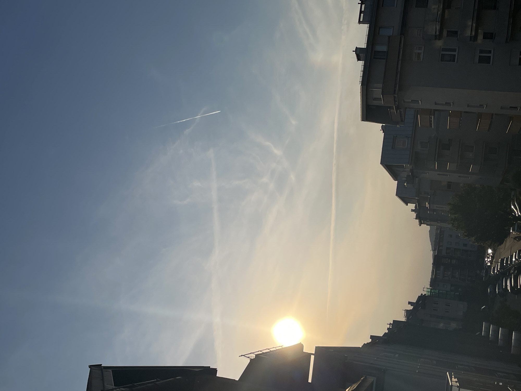 The image displays an urban landscape captured during either sunrise or sunset, evidenced by the sun's low position and the warm, golden light. The sky, occupying the majority of the frame, transitions from a clear, deep blue at the top-left to a hazy, soft yellow and orange near the sun, located at the bottom-center of the rotated original image (which would be the right side of an upright image). The sun is a bright, intense white orb, casting a strong lens flare and partially obscured by the silhouettes of buildings.
Multiple airplane contrails traverse the sky; one prominent, thin white line streaks across the upper-left quadrant, while other more diffused, wispy contrails and light clouds are scattered throughout.
The foreground and right side of the image are dominated by dark silhouettes of multi-story residential or apartment buildings, suggesting a dense urban environment. These structures feature visible windows, balconies, and rooftop details despite being mostly in shadow. Below the sun, the rooftops of buildings are sharply defined against the bright light. In the distance, on the far right, faint outlines of what appear to be hills or mountains blend into the hazy atmosphere behind the buildings. Dark clusters of trees are visible amidst the urban structures. Small, indistinct dark shapes suggestive of parked cars can be seen on a street level between buildings on the far right.
The scene evokes a sense of tranquil transition, blending the natural beauty of the sky with the starkness of an urban cityscape. No visible text is present.