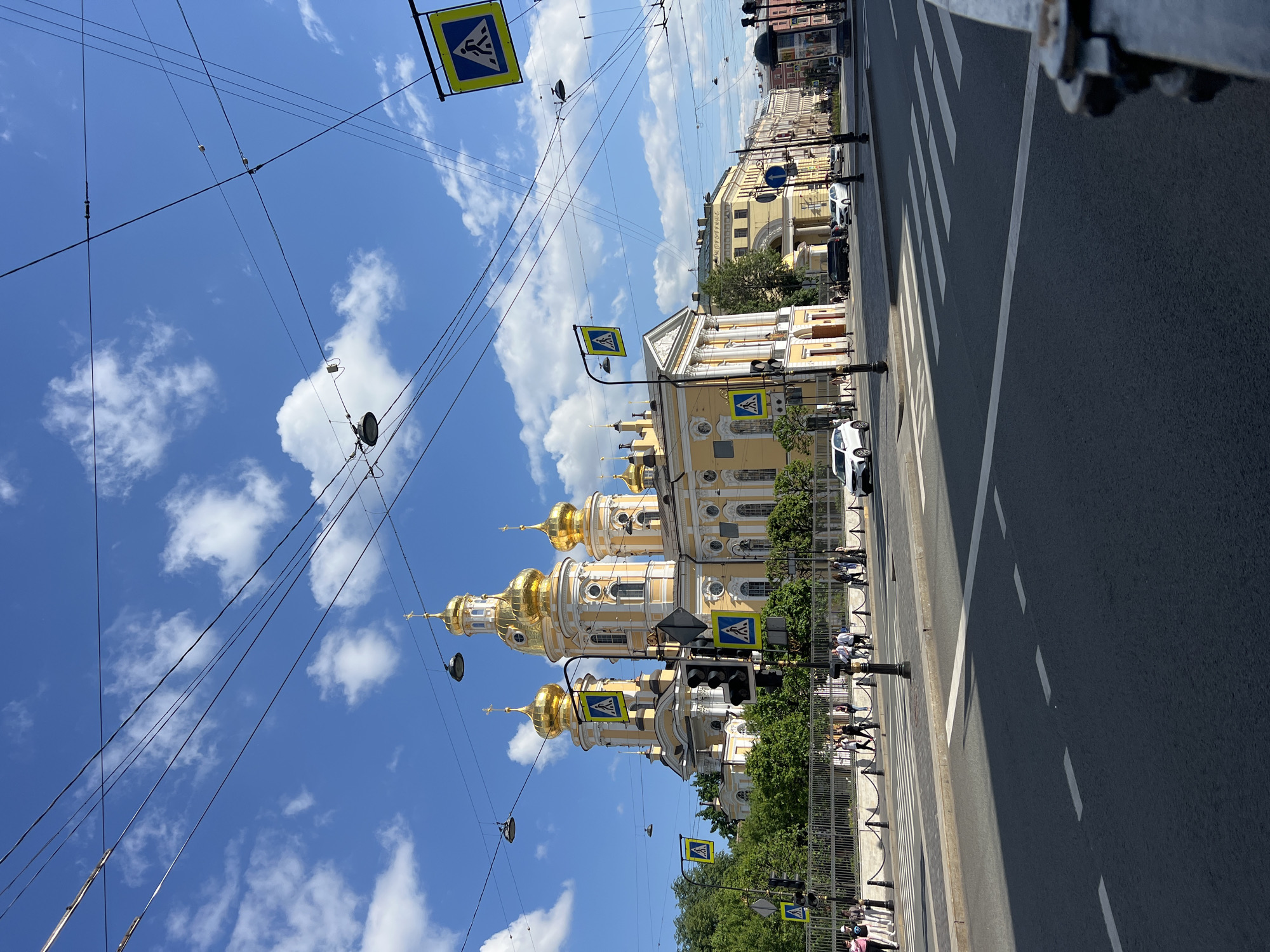 The image captures a view of the Saint Petersburg skyline on a bright, partly cloudy day, with a prominent yellow church adorned with golden domes dominating the scene. The church is located in the city of Saint Petersburg, Russia. The foreground features a street intersection with crosswalks and traffic lights. Overhead, a complex network of electrical wires crisscrosses the sky. Pedestrian crossing signs are visible, and several people are walking on the sidewalks. Cars are present on the road, with one white car stopped at the intersection. The architectural style of the buildings suggests a historical European city. The overall scene conveys a sense of urban activity and architectural grandeur.
