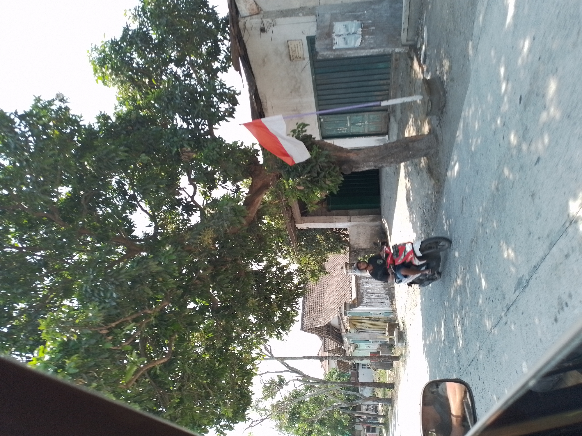 The image depicts a street scene in Wates, Indonesia. In the foreground, the side mirror of a vehicle is visible, reflecting a person's arm. A motorcycle rider, wearing a helmet and a dark t-shirt, is moving away from the viewer on a red and white motorcycle. The rider is on a paved road, which appears to be a local street. To the right of the road, there is a building with a white facade and a green metal gate. A tree with lush green leaves stands in front of the building, and a red and white flag, likely the Indonesian flag, is planted on a pole next to the tree. Behind this building, several houses with terracotta tiled roofs are visible. The environment suggests a rural or semi-urban setting with residential buildings and vegetation. The lighting indicates it is daytime, likely sunny, with shadows cast by trees on the road.