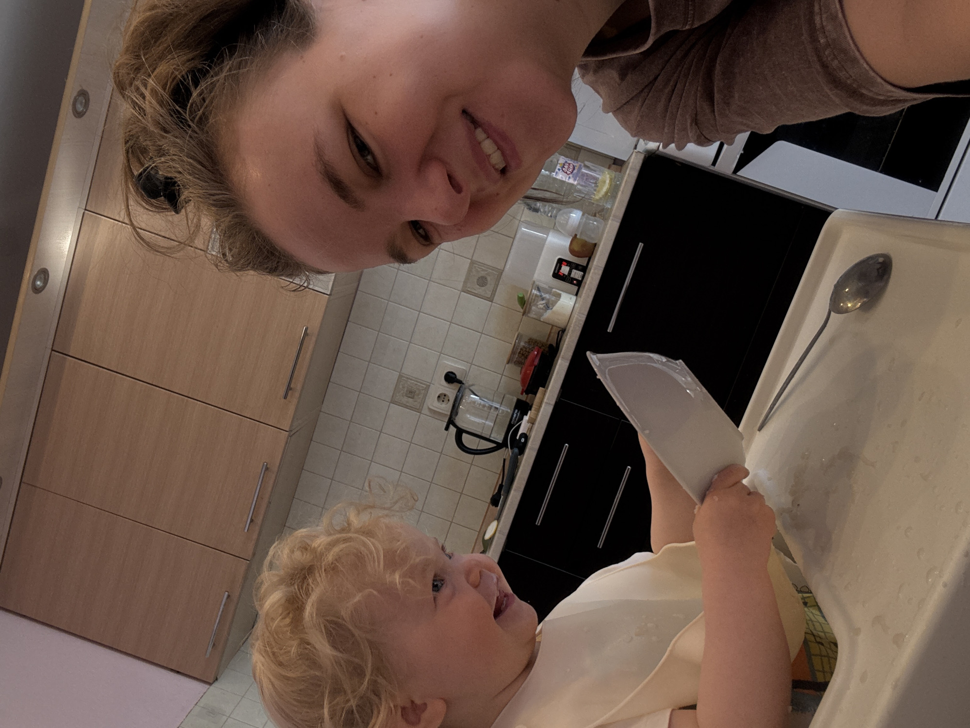 A selfie shows a woman and a young child in what appears to be a kitchen. The woman, with her hair pulled back, is looking into the camera and smiling. The child, with curly blonde hair, is in a high chair, holding a plastic container. A spoon rests on the high chair's tray. The kitchen features light brown cabinets and tiled walls. There's a coffee maker on the countertop.