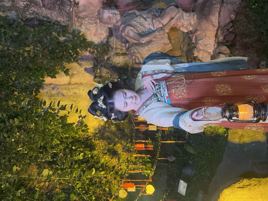 A young East Asian female, positioned slightly right of center, stands in a cultural setting at night within the Hugging Buddha Mountain (抱抱佛山) area of Yuzhong District, Chongqing, China. She is dressed in traditional Hanfu, comprising a light-cream long-sleeved top over a reddish-orange patterned long skirt, accented with a teal sash over her left shoulder and a long white beaded necklace. Her elaborate dark hair is styled with golden hairpins and floral ornaments. Her gaze is directed downwards. She holds a black vintage-style lantern, emitting a warm yellow light, in her extended right hand. Her left hand is placed against her chest. To her left, an ivy-covered wall is visible, adorned with various suspended decorative elements including glowing yellow heart-shaped lights, red rectangular charms, and gold square charms, all hanging from chains. To her right, a light-brown statue depicting a seated child or young monk carrying a large object is partially visible against a textured stone wall. The entire scene is illuminated by artificial yellow lighting.