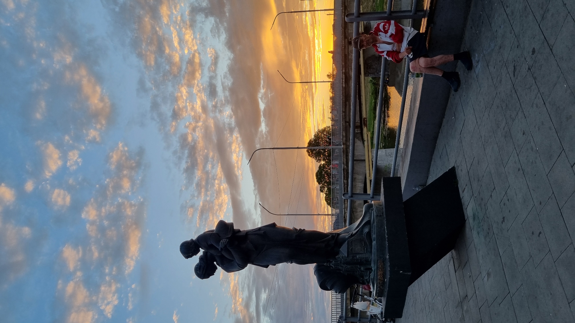 This is a full-frame, eye-level, vertical shot of a monument and a person sitting on a bench near a river at sunset in Mukachevo, Ukraine. The sky is filled with dramatic, orange and pink clouds. In the foreground, a dark bronze sculpture depicts a woman holding a child, with another child or figure at her feet. The sculpture is on a dark, angled pedestal. To the right, a man sits on a bench facing the river. He is wearing a red and white jacket, dark shorts, and black socks and shoes. He is looking towards the left. A bottle of liquid is on the bench next to him. A metal railing runs along the riverbank. Streetlights are visible in the background. The ground is paved with gray rectangular stones. A bridge is visible in the distance over the river.