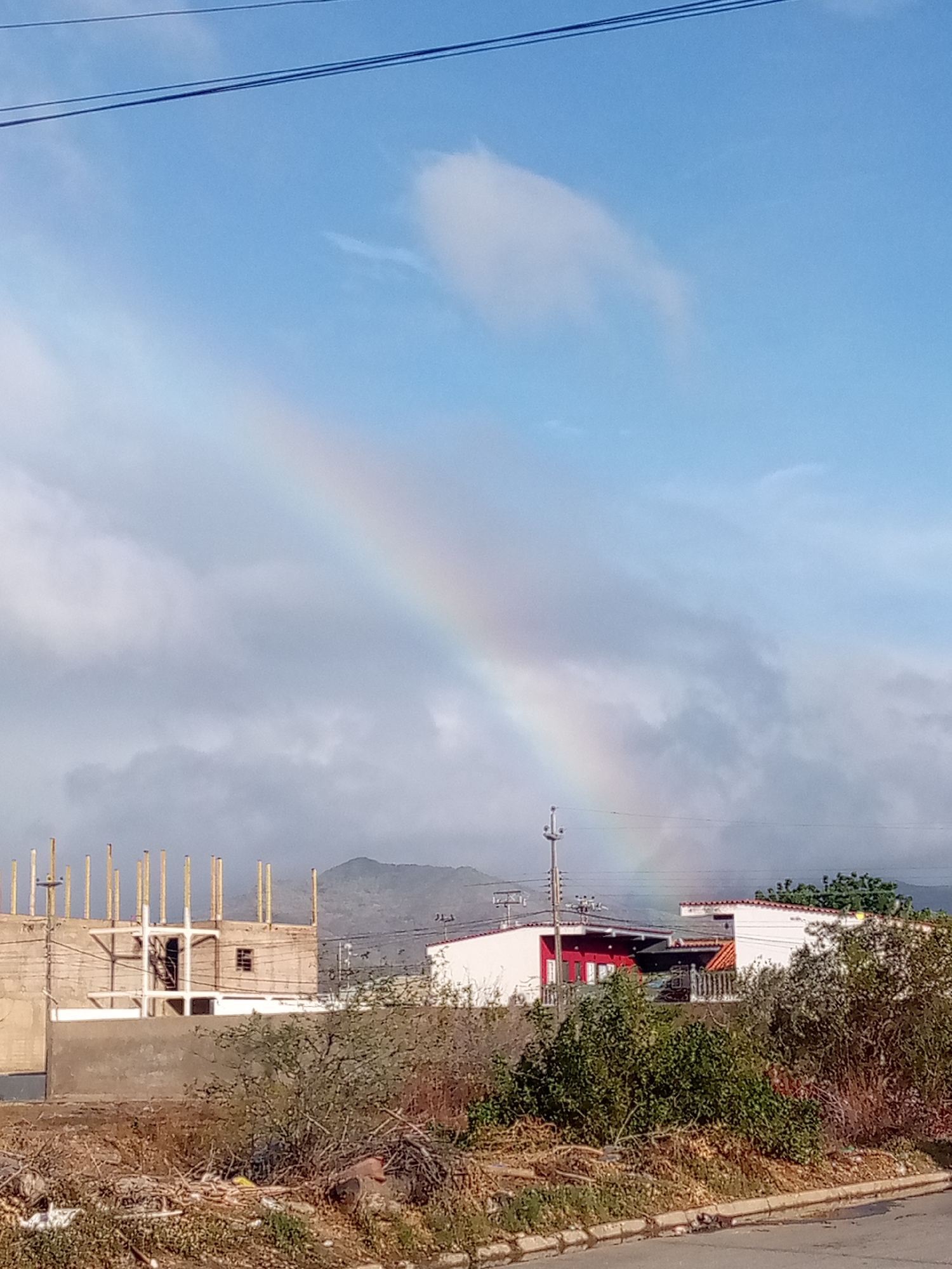 The image captures a daytime scene featuring a rainbow arching across a partly cloudy sky. The sky is predominantly blue with scattered white and gray clouds, and two thin, dark power lines stretch across the top left corner. A faint rainbow is visible, particularly in the upper-left to mid-right sections of the sky, suggesting recent rain or atmospheric moisture.
In the midground, there is a mix of construction and residential buildings. On the left, an unfinished concrete structure with numerous wooden pillars (likely rebar or temporary supports) stands, indicating ongoing construction. To its right, several finished buildings are visible, including a white house with a red facade and a reddish roof-like element, and another white building with an orange-tiled roof further behind it. A utility pole with wires is also present in this section.
The foreground is characterized by dry, sparse vegetation, including overgrown bushes and what appears to be discarded debris or dry brush on the ground. A gray curb and a section of paved road are visible at the very bottom right. In the distant background, hazy mountains rise under the arc of the rainbow.
The overall impression is that of a semi-urban or developing area, possibly in a region prone to dry periods, with elements of nature and human activity coexisting. There is no visible text in the image.