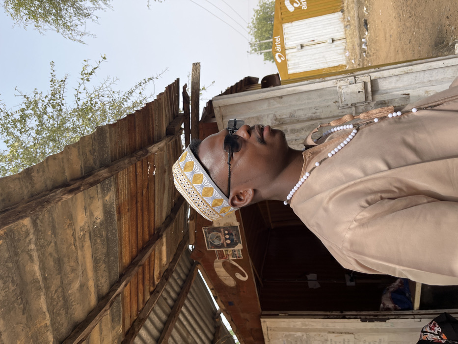 The image depicts an outdoor scene in Kanti, Nigeria, under bright daylight. A young adult male is positioned in the foreground, seen in profile, looking towards the right. He possesses a medium-dark complexion, wears dark sunglasses, a light brown traditional garment, and a white and gold embroidered kufi cap. A long white beaded necklace with two tassels is draped around his neck. Behind the individual, to the left, is a weathered, small wooden structure, likely a stall or shop, featuring a rusty corrugated metal roof supported by wooden beams. Faded posters are visible on the structure's wall, and an open doorway reveals a dimly lit interior. To the right in the background, a yellow structure, possibly a gate or kiosk, displays the red "Airtel" logo. The ground is unpaved earth, and sparse tree foliage is visible against a clear blue sky.