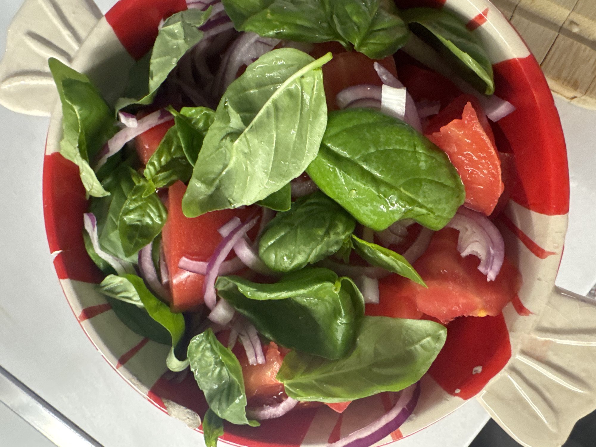 A close-up shot shows a bowl of tomato and basil salad. The bowl is red and white striped with a decorative rim. Sliced red onions are mixed with chopped tomatoes and fresh green basil leaves. The salad appears to be on a white surface, possibly a table, with a metallic object and a hint of a wooden surface in the background. The image does not provide any information about the location in Sofia, Bulgaria.