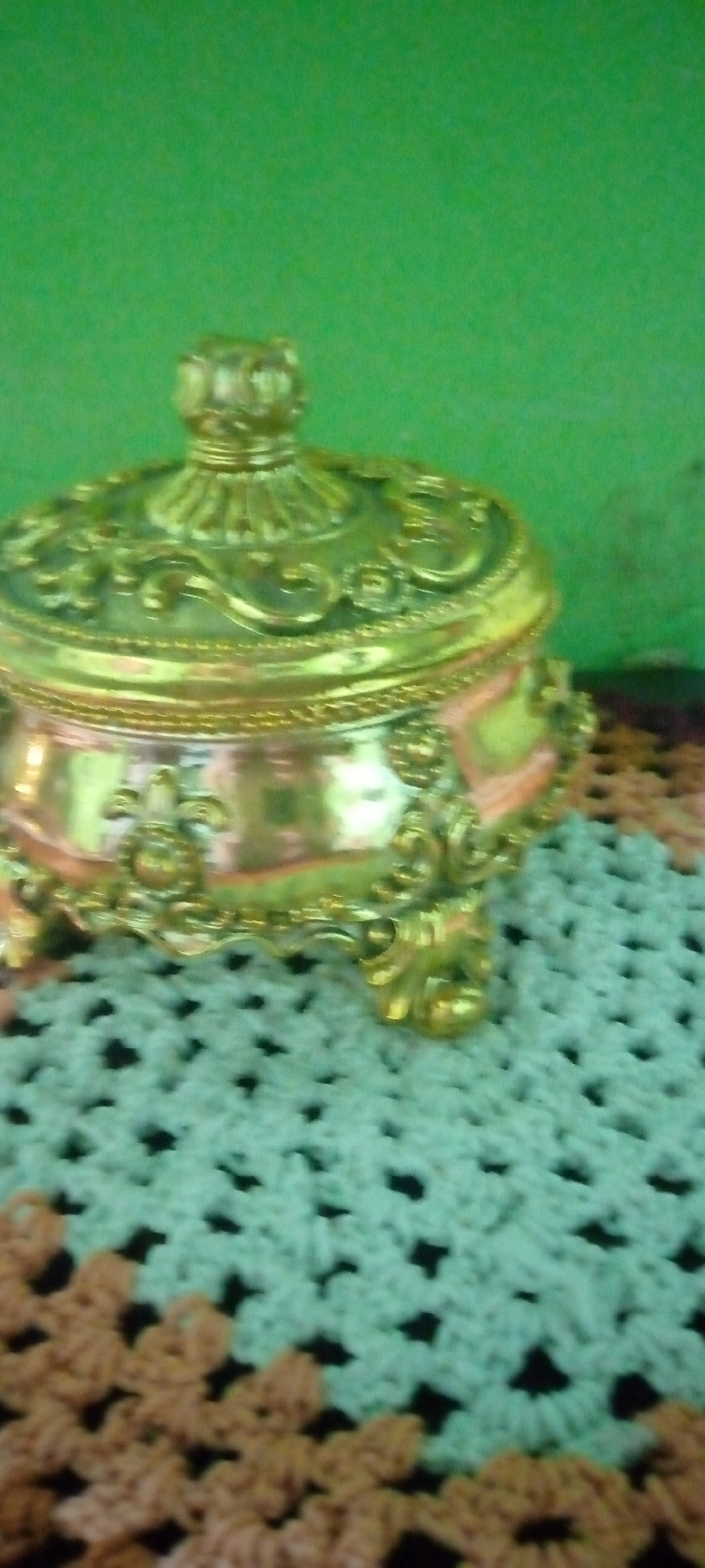 An ornate, gold-colored decorative box with intricate detailing sits on a crocheted surface. The box has a lid with a raised, sculpted design and rests on three decorative feet. The crocheted surface is made of alternating light green and dark brown squares, creating a patterned texture. The background is a solid, bright green wall. The image does not contain any identifiable landmarks or clues to determine its location in Sumberwono, Indonesia.