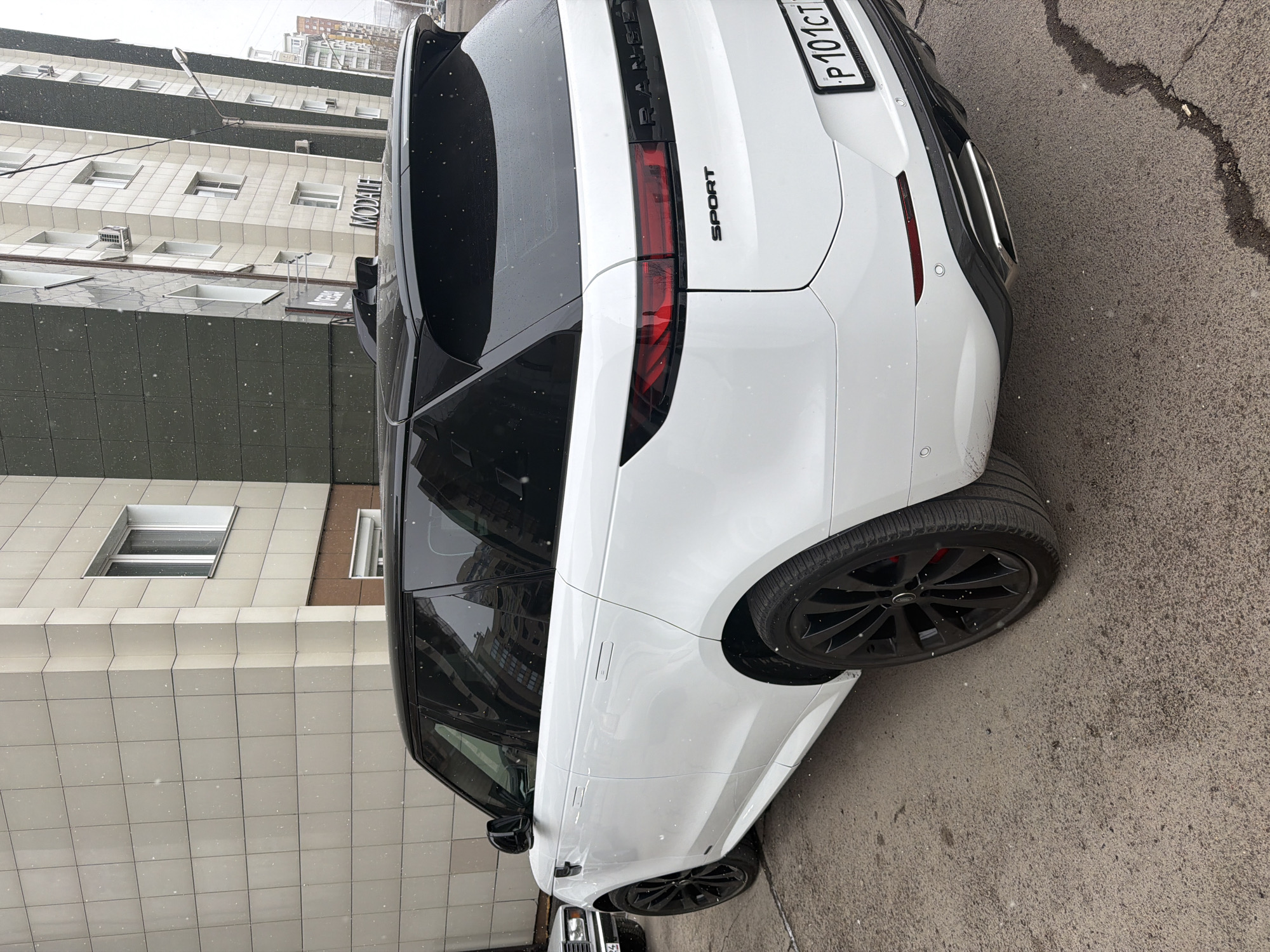The image shows the rear of a white Range Rover Sport parked on a street, likely in the city of Krasnoyarsk, Russia, based on the license plate which reads "P 101 CT". The vehicle is parked next to a building with a sign that reads "МОДАLI". Snow is falling. The ground is asphalt.