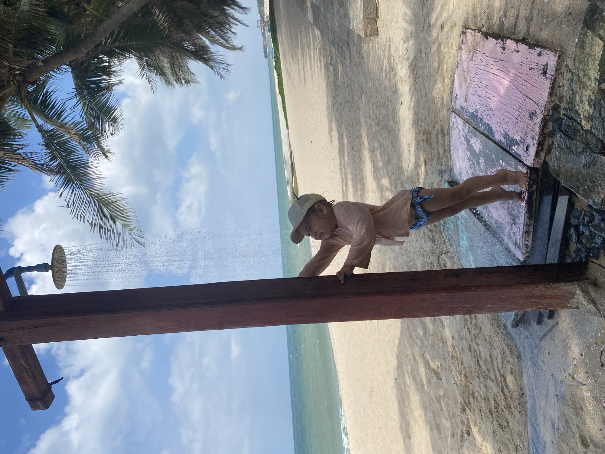 A young child, wearing a light peach-colored long-sleeved shirt and blue-patterned swim trunks, stands on a pink wooden platform by a beach shower. The child is holding onto the wooden post of the shower, looking up at the water spray. The beach has light-colored sand, and the ocean is visible in the background, with gentle waves. Palm trees are present, with their fronds casting shadows on the sand. The sky is blue with scattered white clouds. The location is identified as Phu Thuy Ward, Vietnam.